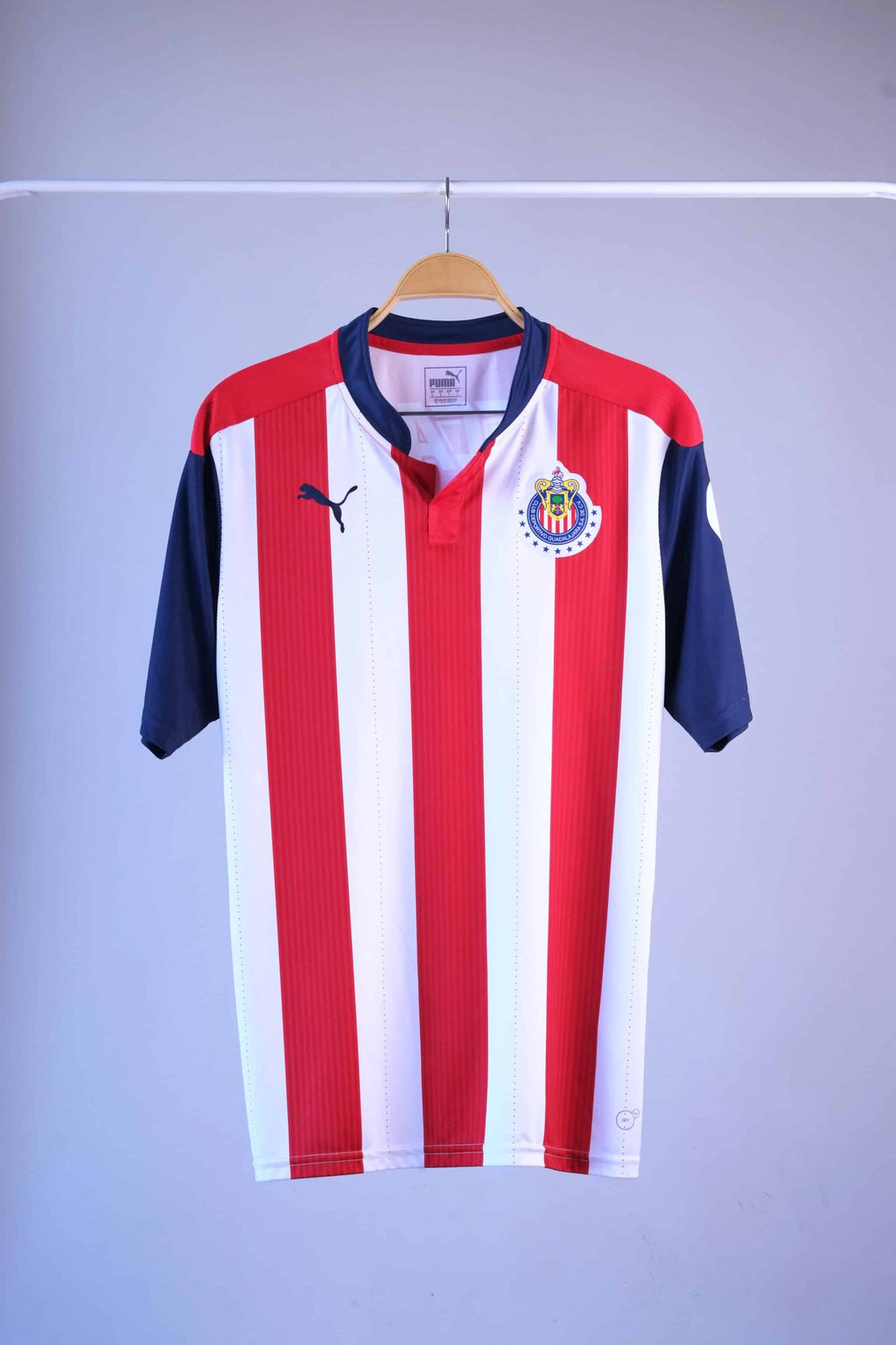 Front view of a white and red vertically striped football jersey hanging on a wooden hanger. It features short navy blue sleeves, a navy blue collar with a small buttonless Henley-style placket, a blue Puma logo on the right breast, and the circular Chivas club crest on the left breast.