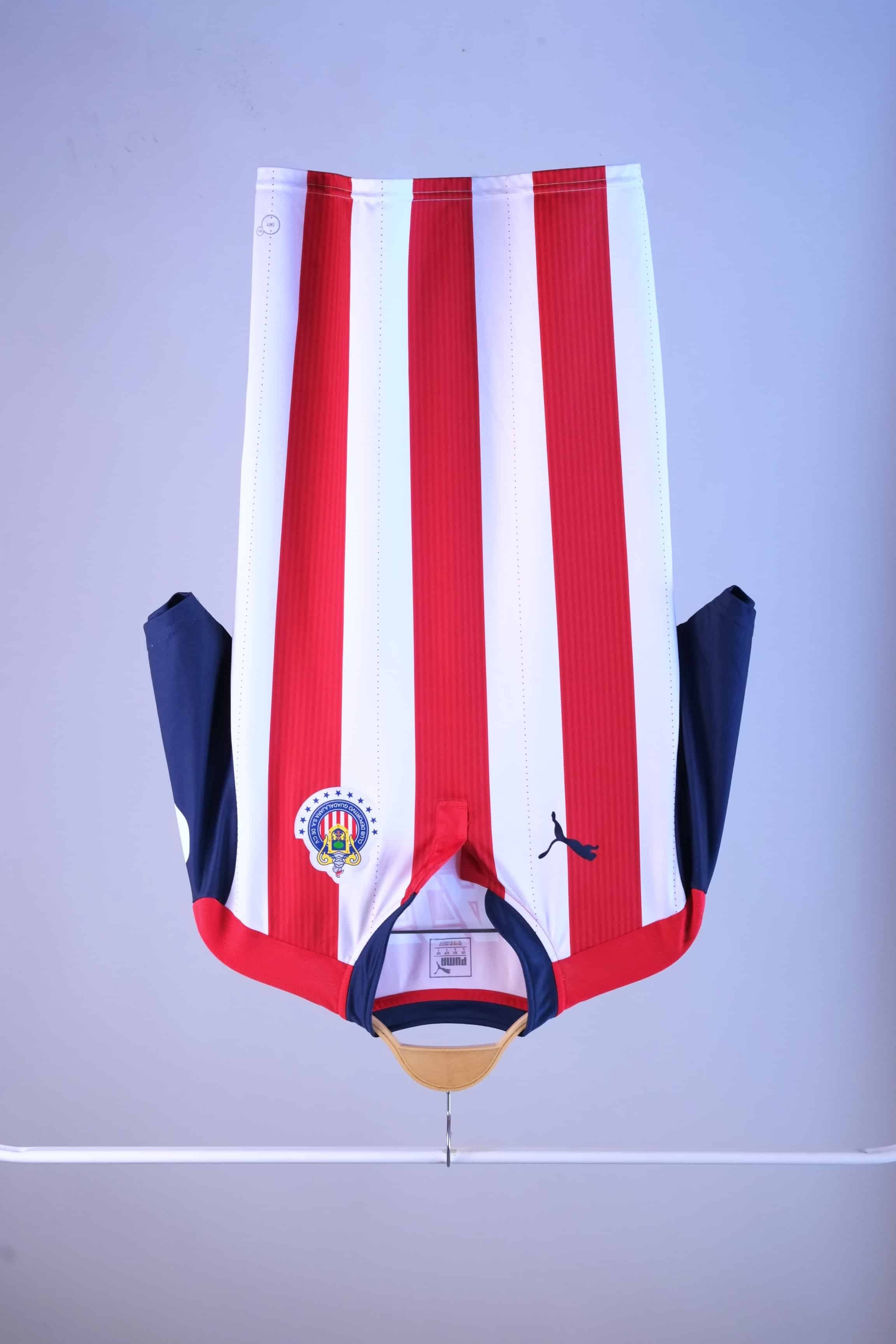 Front view of a white and red vertically striped football jersey hanging on a wooden hanger. It features short navy blue sleeves, a navy blue collar with a small buttonless Henley-style placket, a blue Puma logo on the right breast, and the circular Chivas club crest on the left breast.