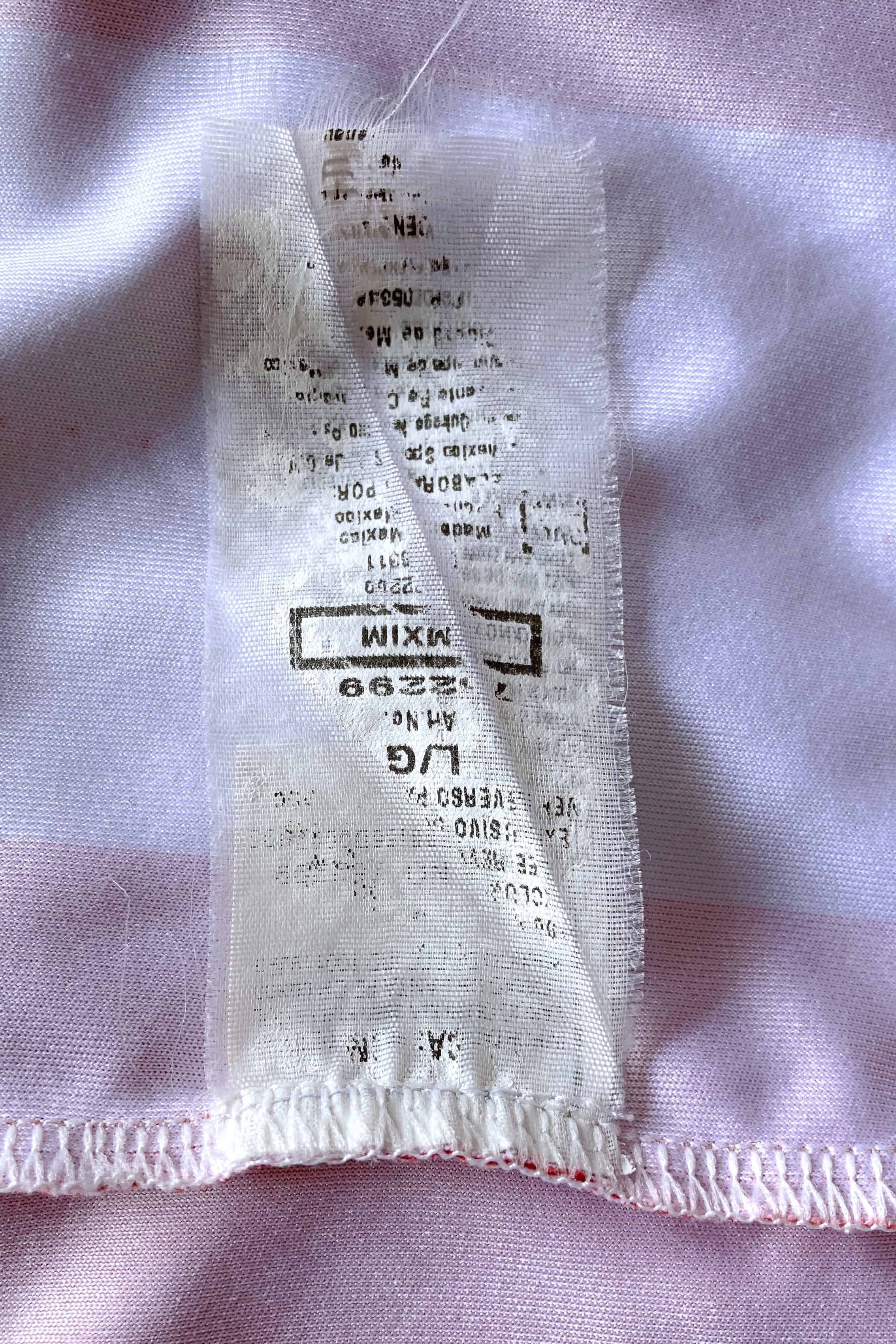 Close-up of a long, semi-translucent white fabric care label stitched inside the shirt against a red and white striped backdrop. The tag displays printing that includes the sizing "L/G", "Art No. 762299", "MXIM", and manufacturing details indicating it was made in Mexico.