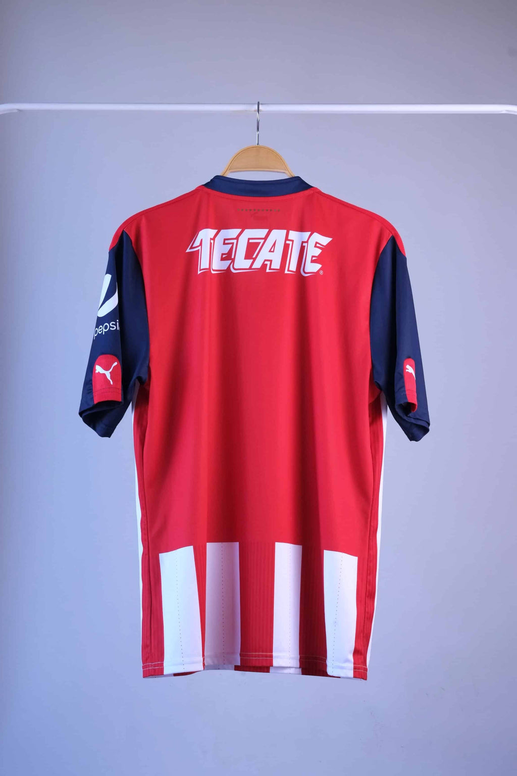 Back view of the striped football jersey on a hanger. The back is predominantly red, featuring a large white "TECATE" sponsor logo across the shoulders and a white and red striped pattern extending along the lower hem.