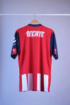 Back view of the striped football jersey on a hanger. The back is predominantly red, featuring a large white "TECATE" sponsor logo across the shoulders and a white and red striped pattern extending along the lower hem.