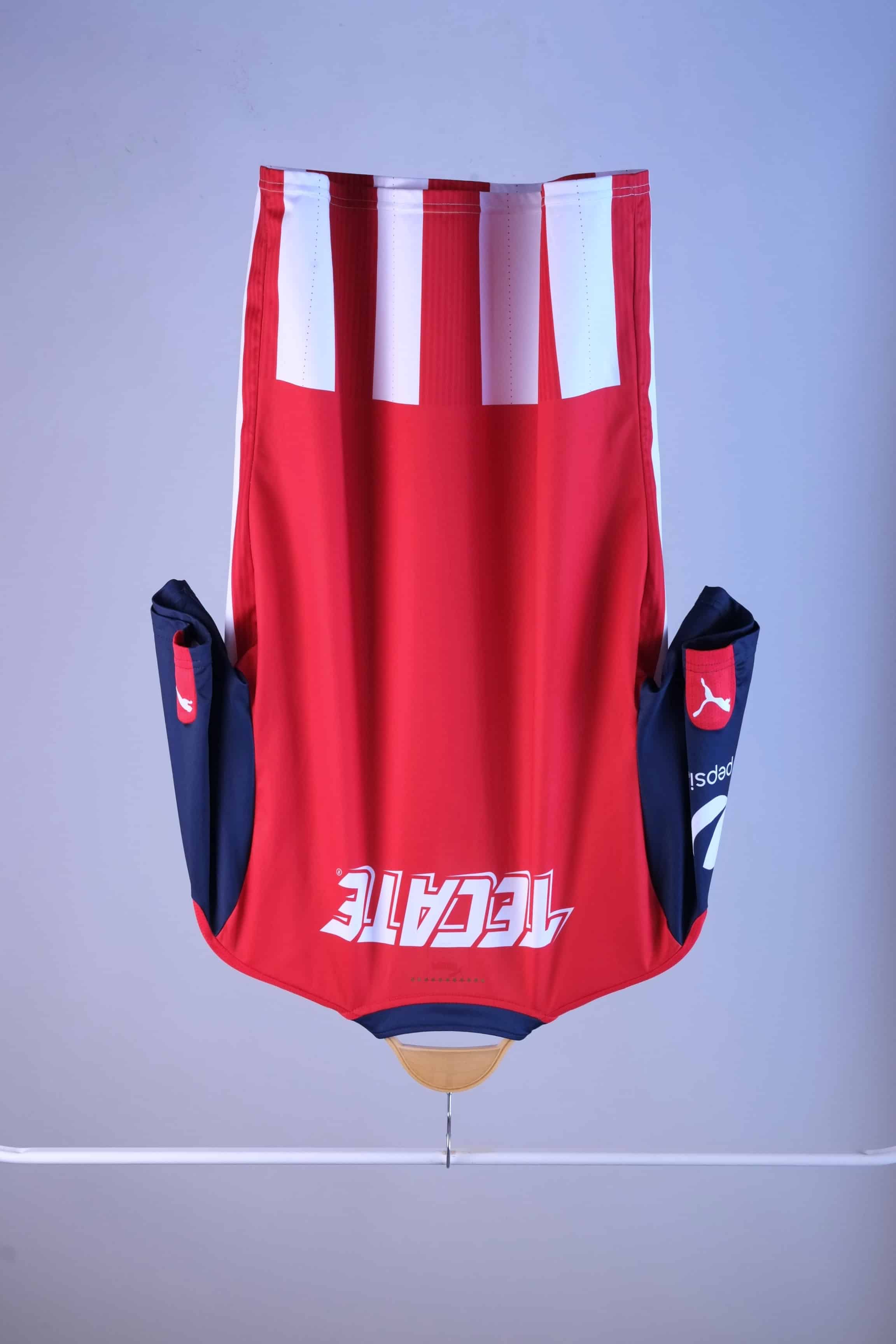 Back view of the striped football jersey on a hanger. The back is predominantly red, featuring a large white "TECATE" sponsor logo across the shoulders and a white and red striped pattern extending along the lower hem.