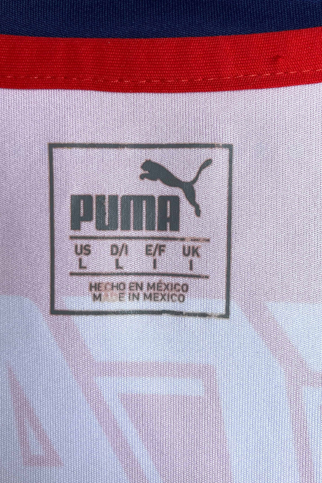 Close-up of the square, heat-pressed size label located on the inner neck. It features the classic Puma logo, text reading "US L / D/I L / E/F L / UK L", and "HECHO EN MEXICO / MADE IN MEXICO" stamped below the sizing grid against the white jersey fabric.