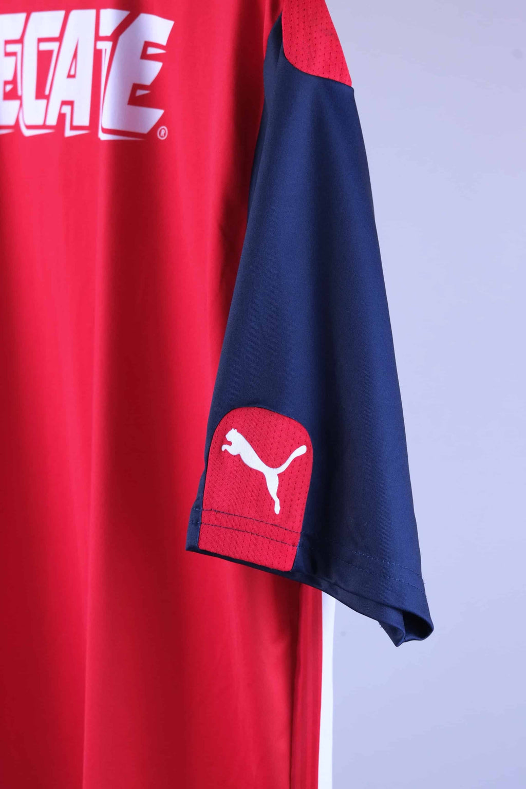 Detailed close-up view of the jersey's navy blue sleeve showcasing a red panel at the cuff containing a white Puma cat logo. The solid red torso of the shirt and a section of the white "TECATE" back graphics are visible to the left.
