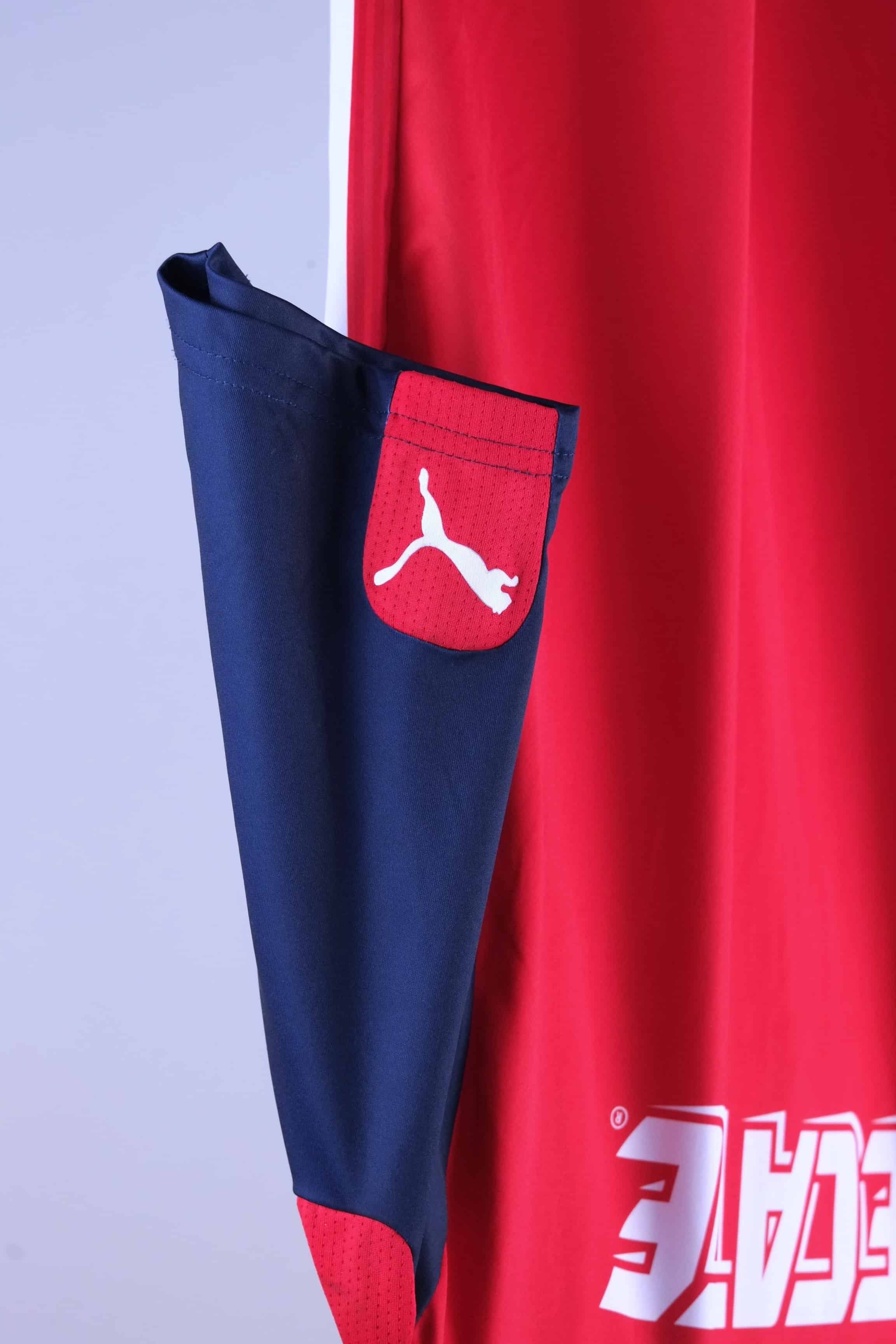 Detailed close-up view of the jersey's navy blue sleeve showcasing a red panel at the cuff containing a white Puma cat logo. The solid red torso of the shirt and a section of the white "TECATE" back graphics are visible to the left.