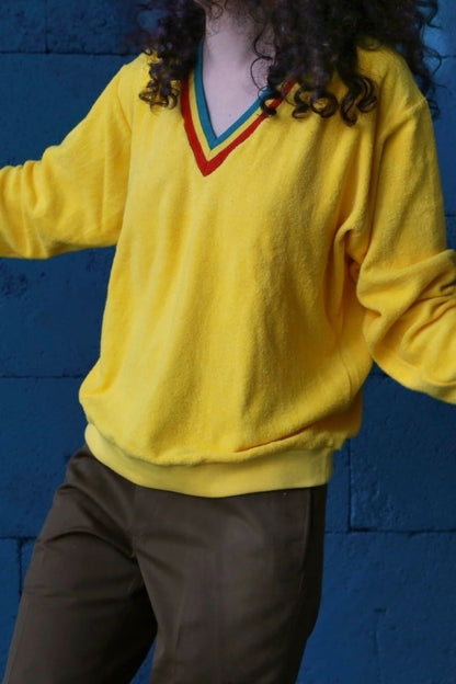 Person wearing a vintage 70s yellow terrycloth V-neck sweater with a colorful striped collar in red, green, and blue, styled casually against a dark blue background.