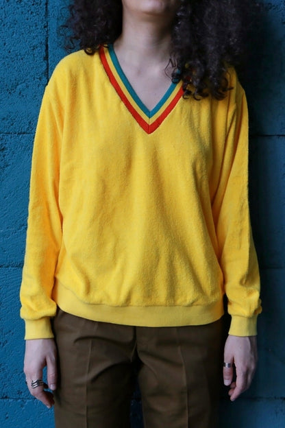 Person with curly dark hair wearing a vintage 70s yellow terrycloth V-neck sweater with a colorful striped collar in red, green, and blue, standing against a dark blue background.