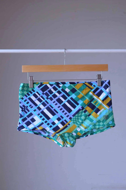 Ribana vintage 70s geometric swim trunks in blue and green with abstract check pattern, hanging on wooden hanger.