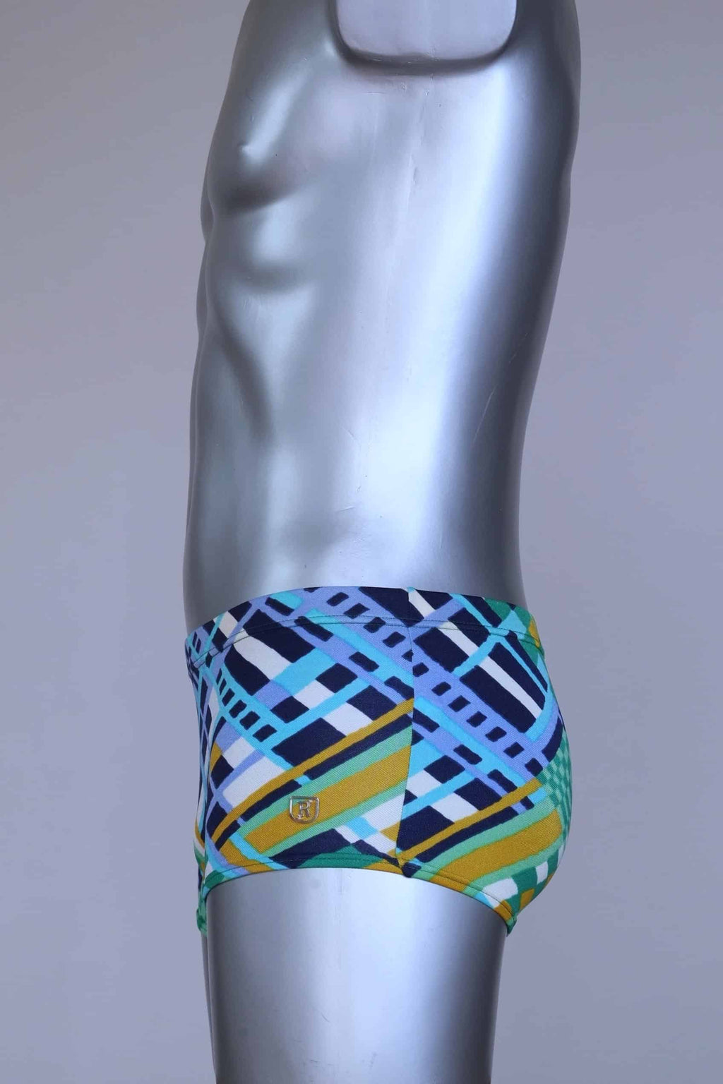 Side view of vintage 70s Ribana swim trunks in a geometric print with cool tones of blue, green, yellow, and navy, modeled on a silver mannequin.