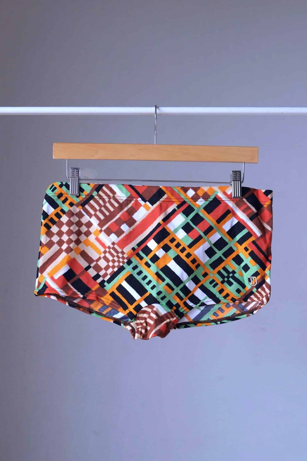 Ribana vintage 70s geometric swim trunks in orange and brown with abstract check pattern, hanging on wooden hanger.