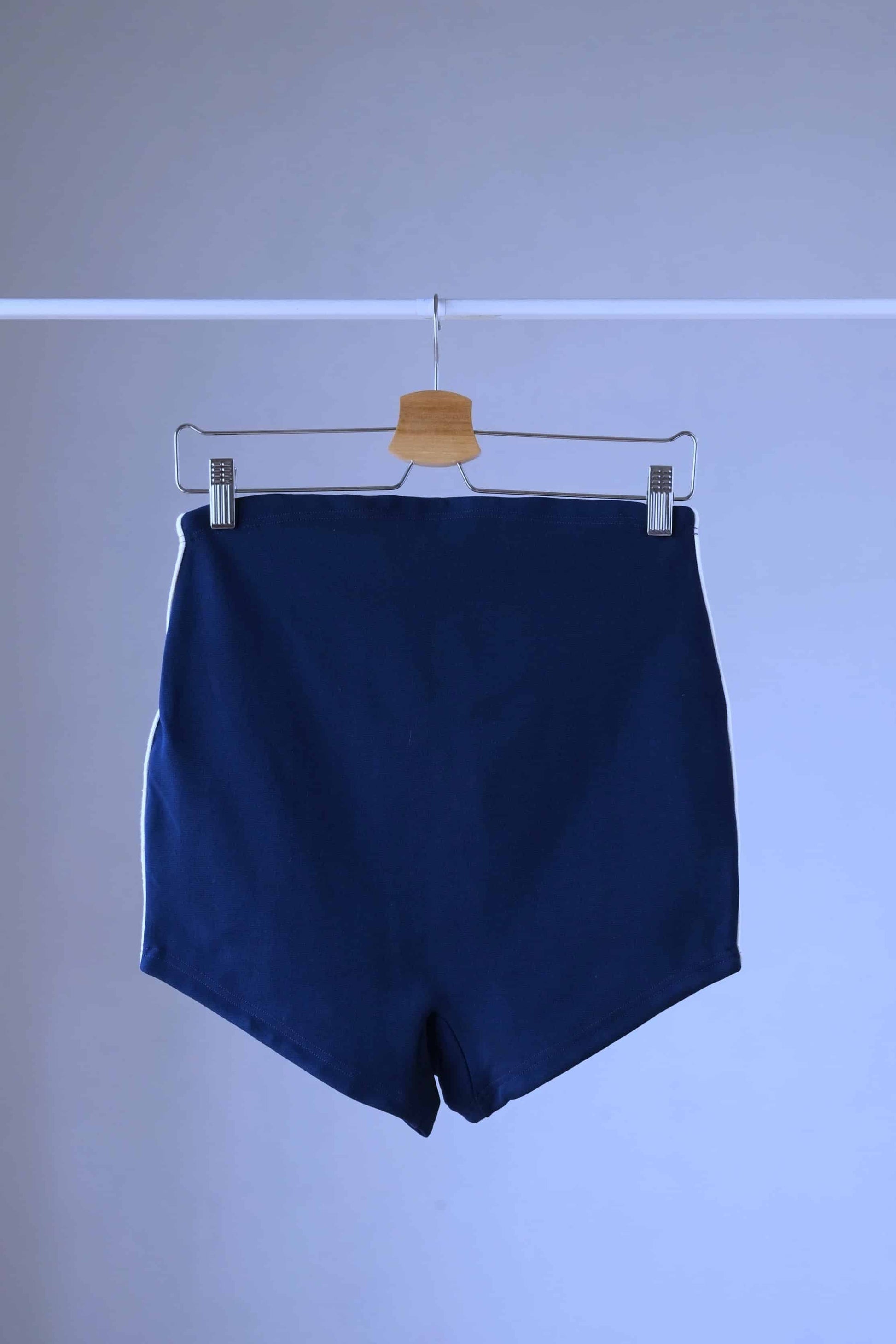 Back shot of A pair of Ribana Oscar 1950's vintage high-waisted navy blue swim trunks with white piping along the sides, hanging on a hanger against a plain background