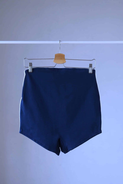 Back shot of A pair of Ribana Oscar 1950's vintage high-waisted navy blue swim trunks with white piping along the sides, hanging on a hanger against a plain background