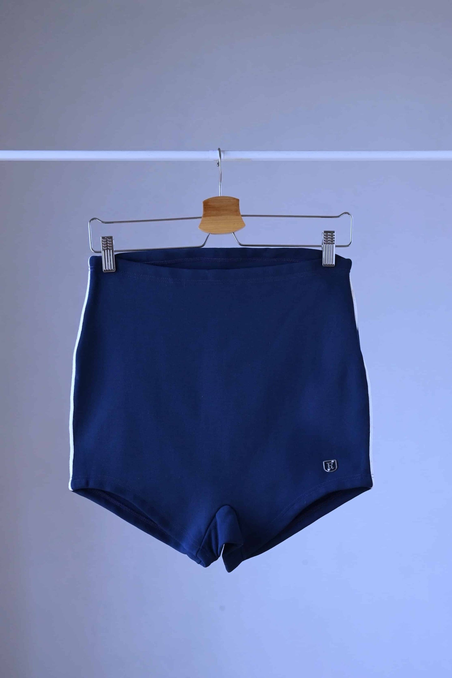 A pair of Ribana Oscar 1950's vintage high-waisted navy blue swim trunks with white piping along the sides, hanging on a hanger against a plain background