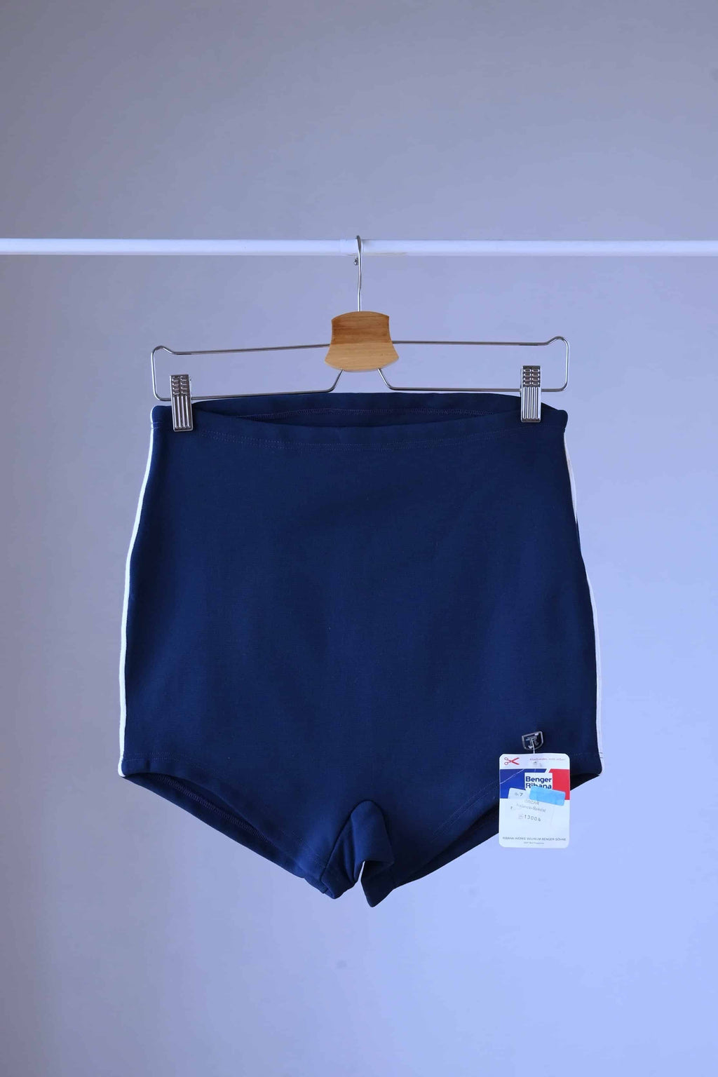 A pair of Ribana Oscar 1950's vintage high-waisted navy blue swim trunks with white piping along the sides, hanging on a hanger against a plain background with their tag showing