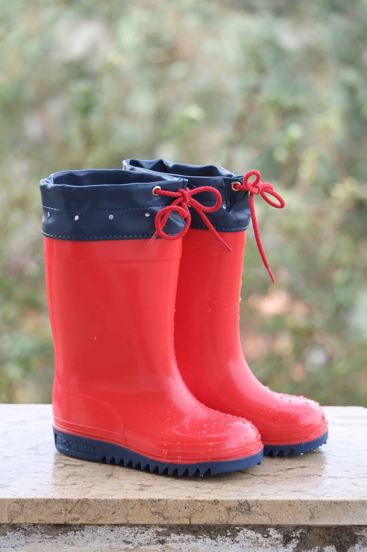 Red and navy ROMIKA Bobby Rain Boots on a ledge with raindrops on the boots