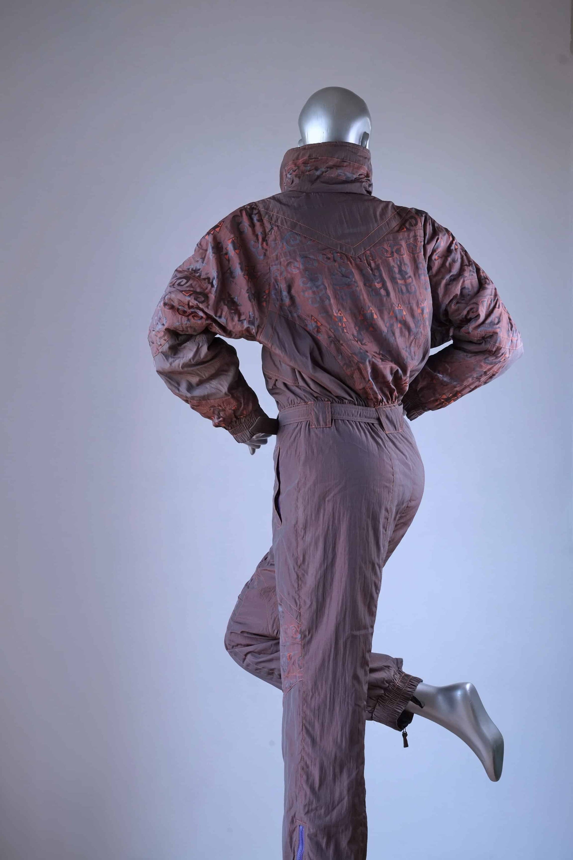 back view of a brown vintage 90s ski suit with a subtle orange pattern, shown on a mannequin with one leg raised.