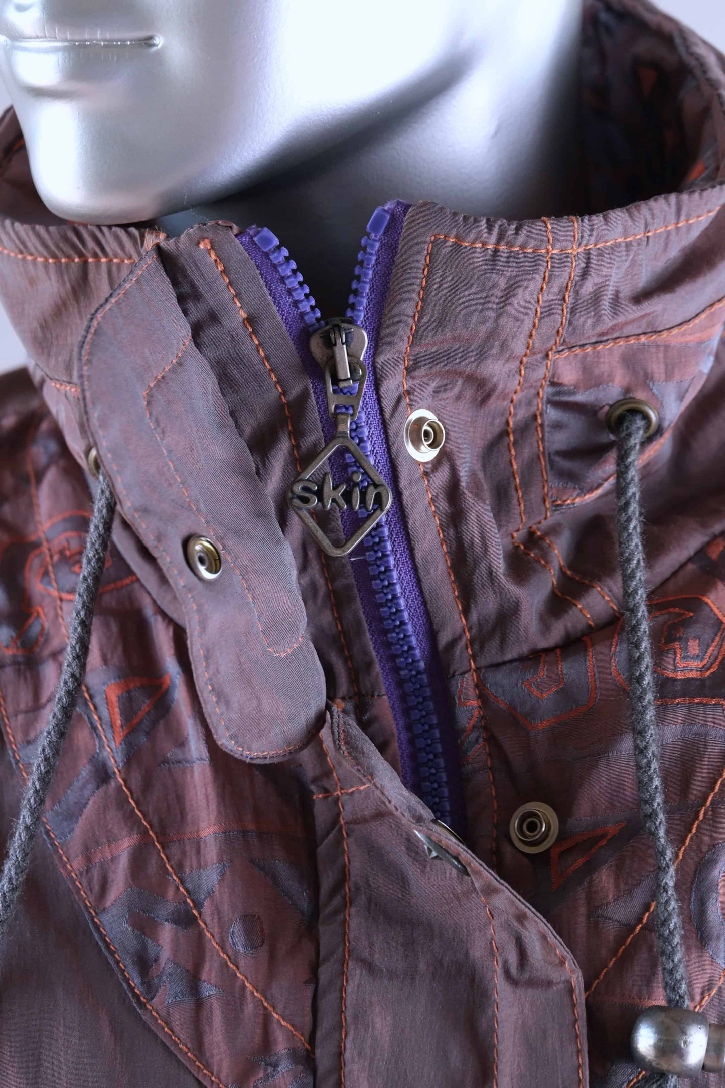 Detailed view of a brown 90s ski suit collar, showing a purple zipper, snap closures, drawstrings, and a 'Skin' brand zipper pull.