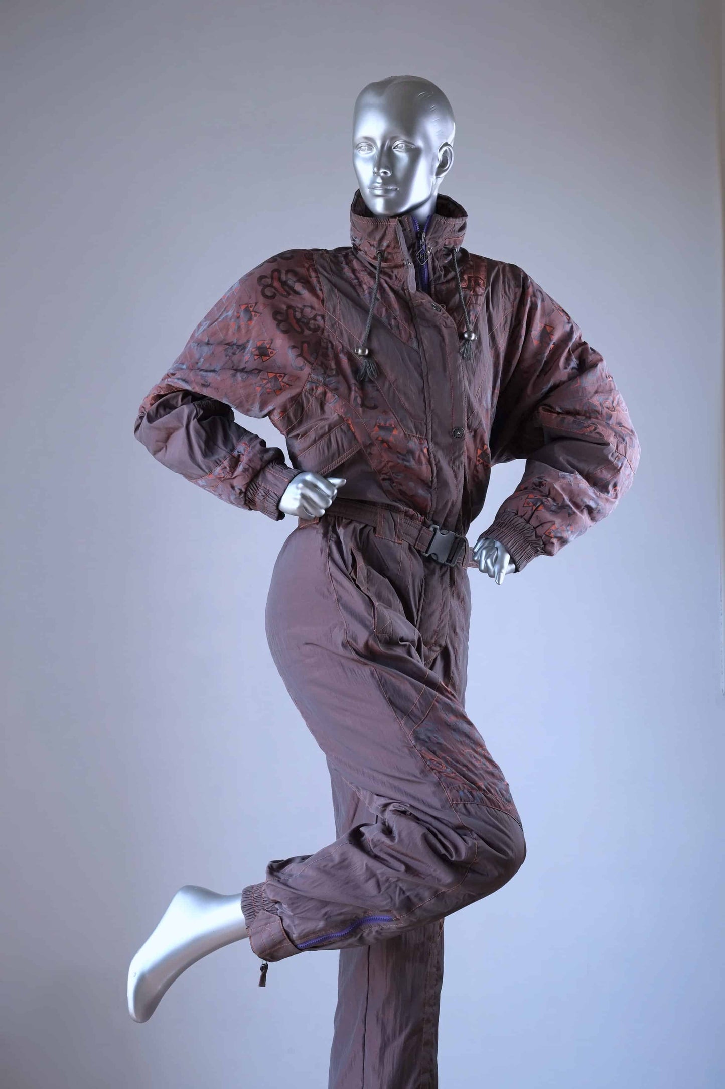 A brown vintage 90s ski suit with a subtle orange pattern, shown on a mannequin with one leg raised.