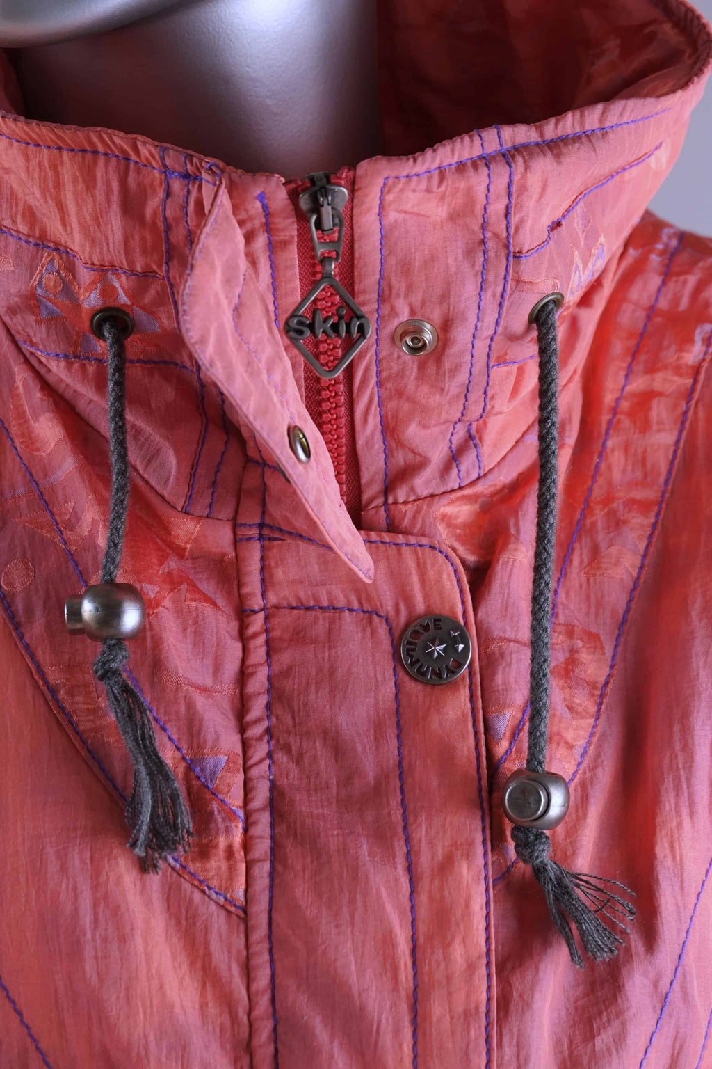 Detailed view of a coral 90s ski suit collar, showing a zipper, snap closures, drawstrings, and a 'Skin' brand zipper pull.