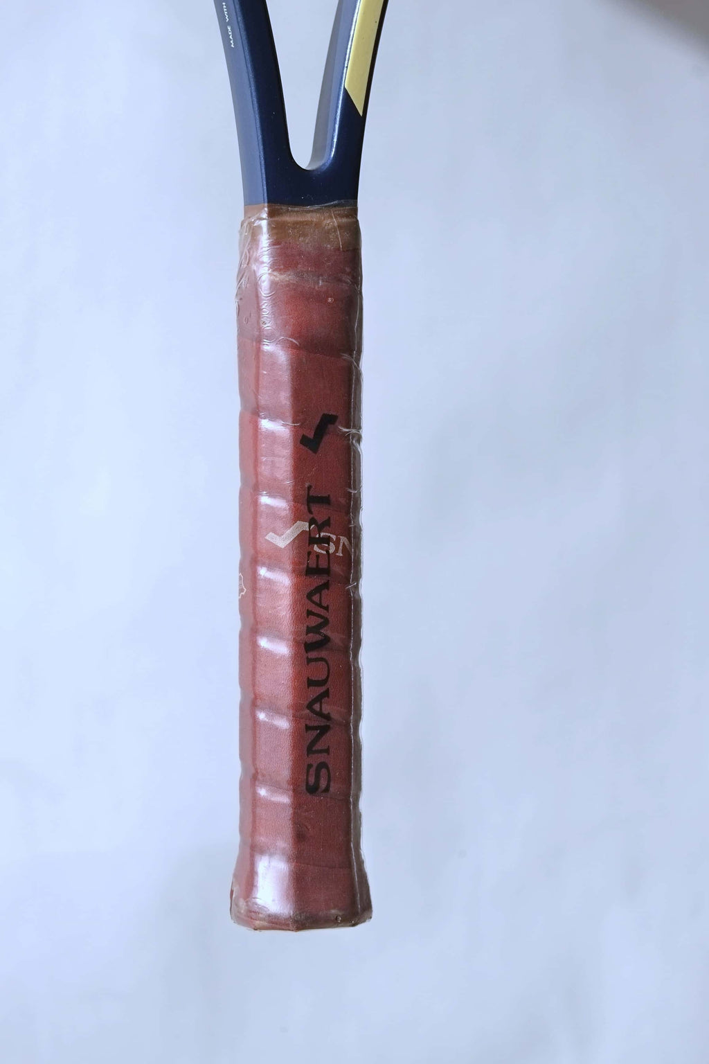 Detail shot of the racquet handle wrapped in its original brown leather Snauwaert-branded grip, encased in a clear protective plastic seal.