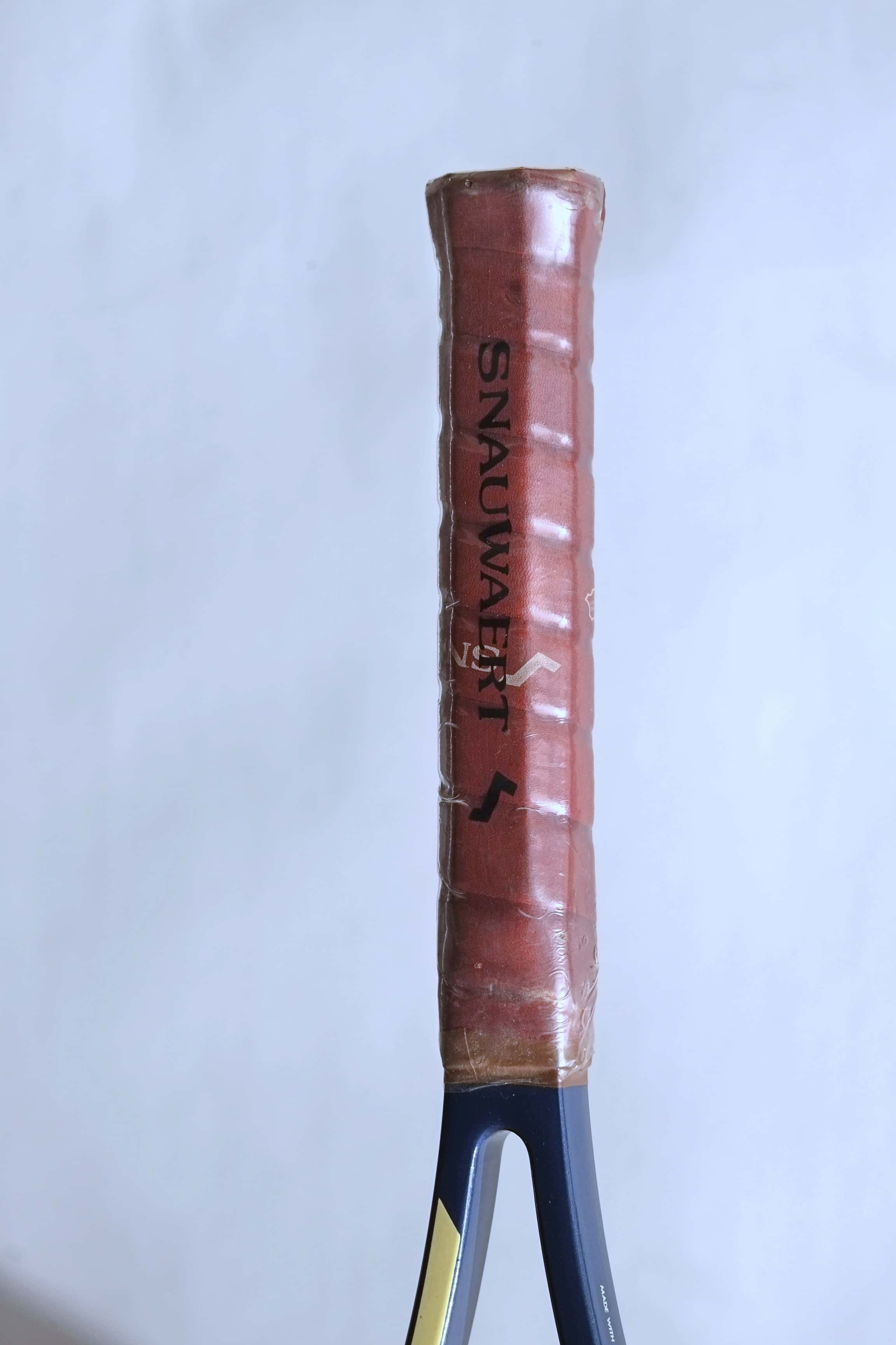 Detail shot of the racquet handle wrapped in its original brown leather Snauwaert-branded grip, encased in a clear protective plastic seal.