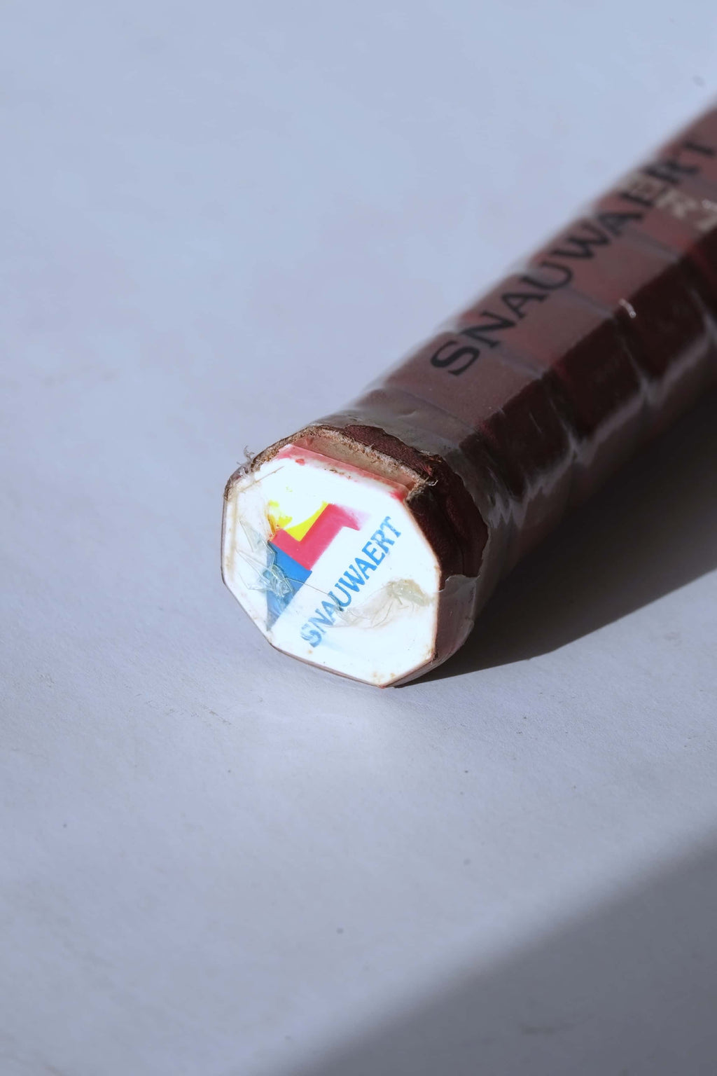 Angled close-up of the white racquet butt cap featuring the colorful Snauwaert geometric logo and name, sealed in original plastic film over a brown leather grip.