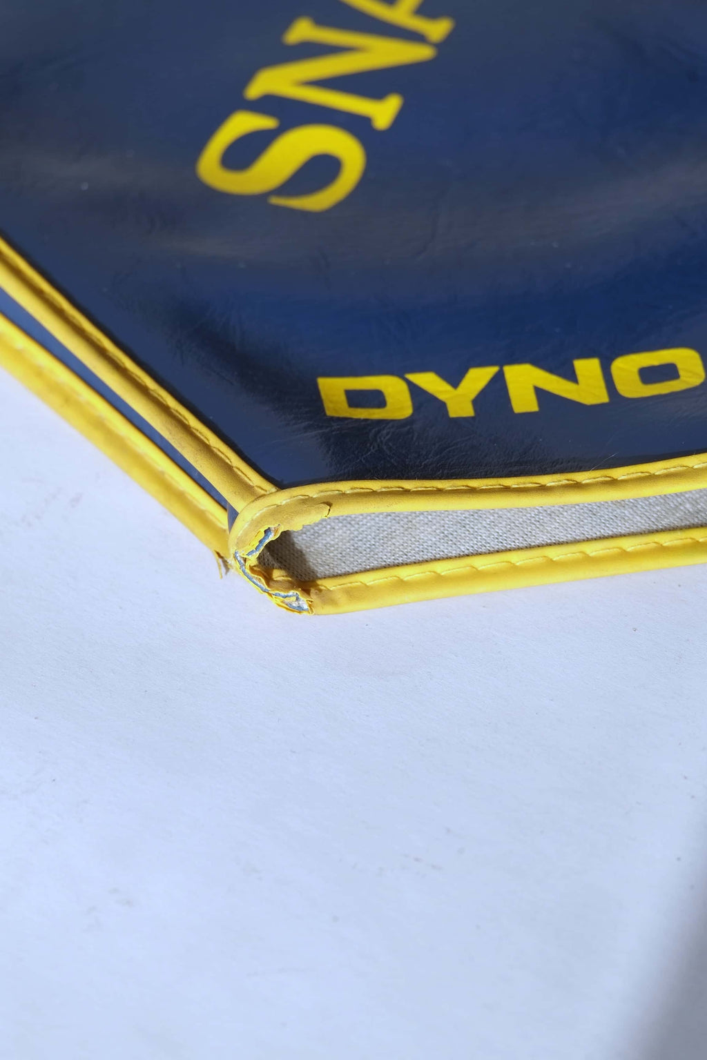 Extreme close-up of the stitched yellow trim on a navy blue vinyl tennis racquet cover, showing slight vintage wear at the corner.