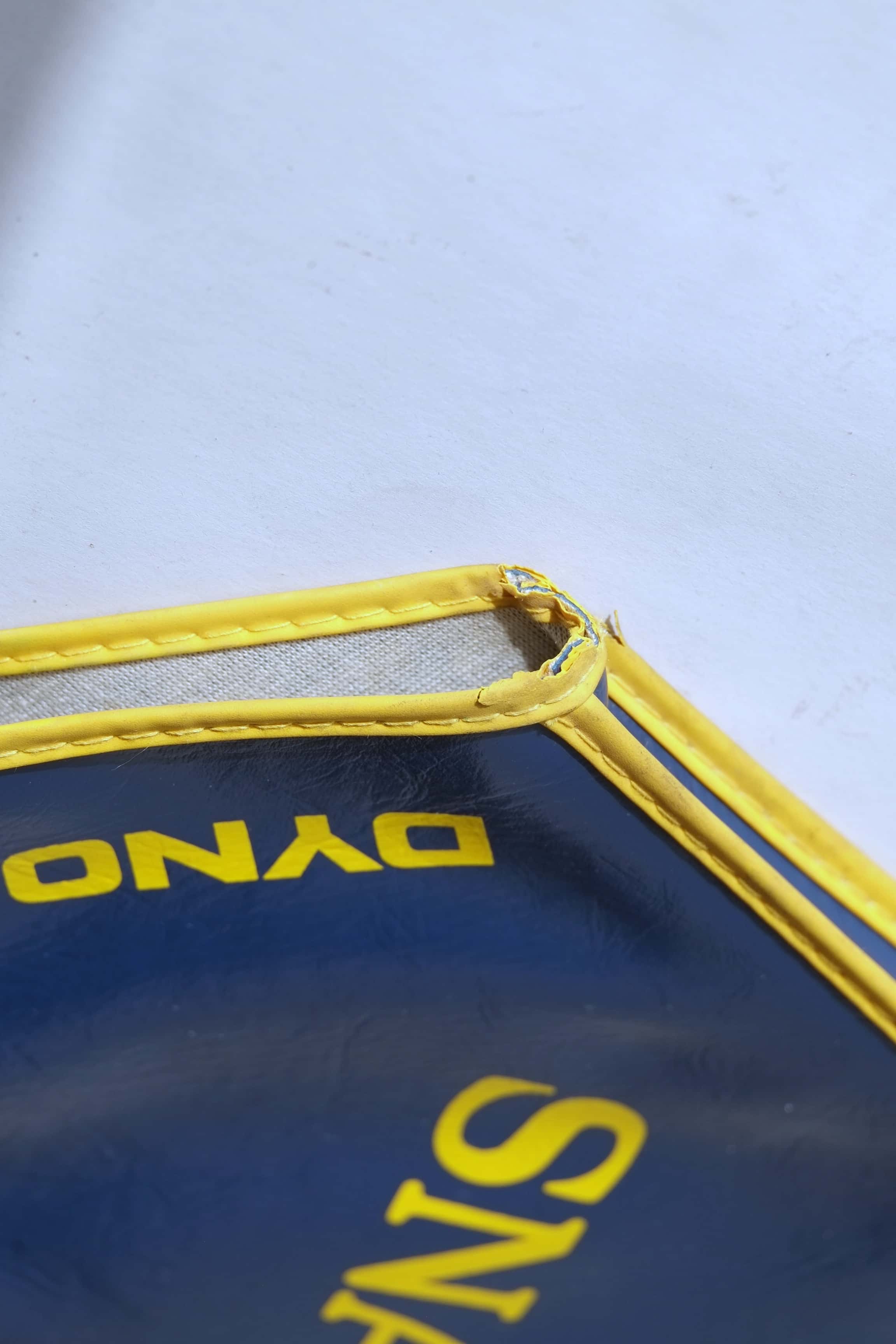 Extreme close-up of the stitched yellow trim on a navy blue vinyl tennis racquet cover, showing slight vintage wear at the corner.