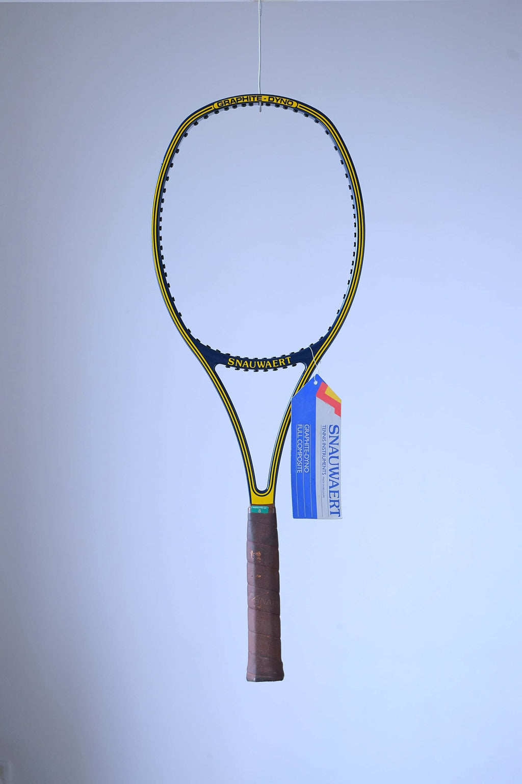 Full studio shot of an unstrung vintage Snauwaert Graphite Dyno tennis racquet hanging vertically, showing its blue and yellow frame, brown leather grip, and a large blue and white paper hang tag attached to the throat.