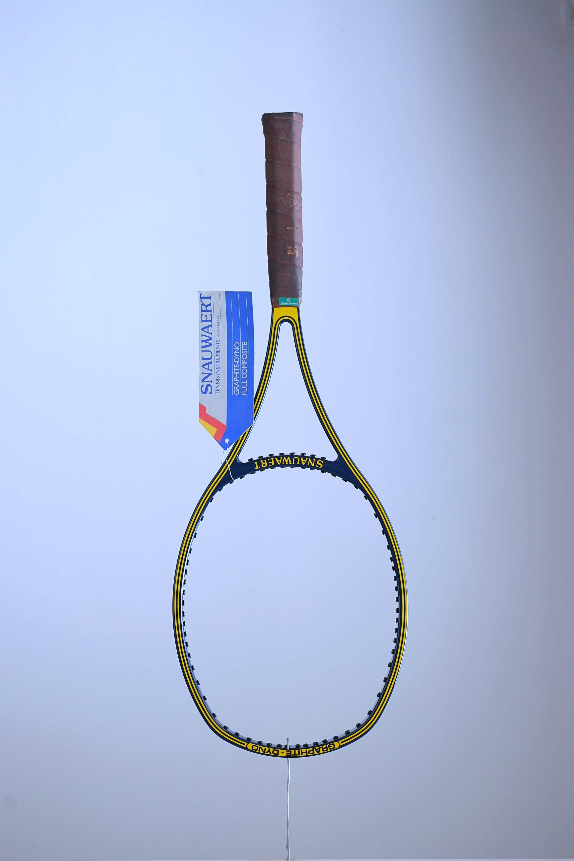 Full studio shot of an unstrung vintage Snauwaert Graphite Dyno tennis racquet hanging vertically, showing its blue and yellow frame, brown leather grip, and a large blue and white paper hang tag attached to the throat.