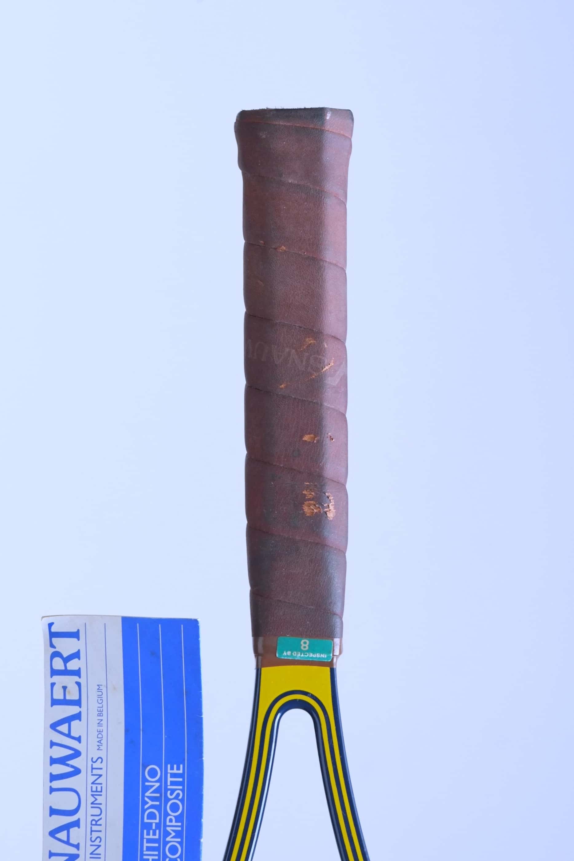 Close-up of the racquet's original brown leather grip showing minor surface wear, a green inspection sticker at the top collar, and the attached paper hang tag.