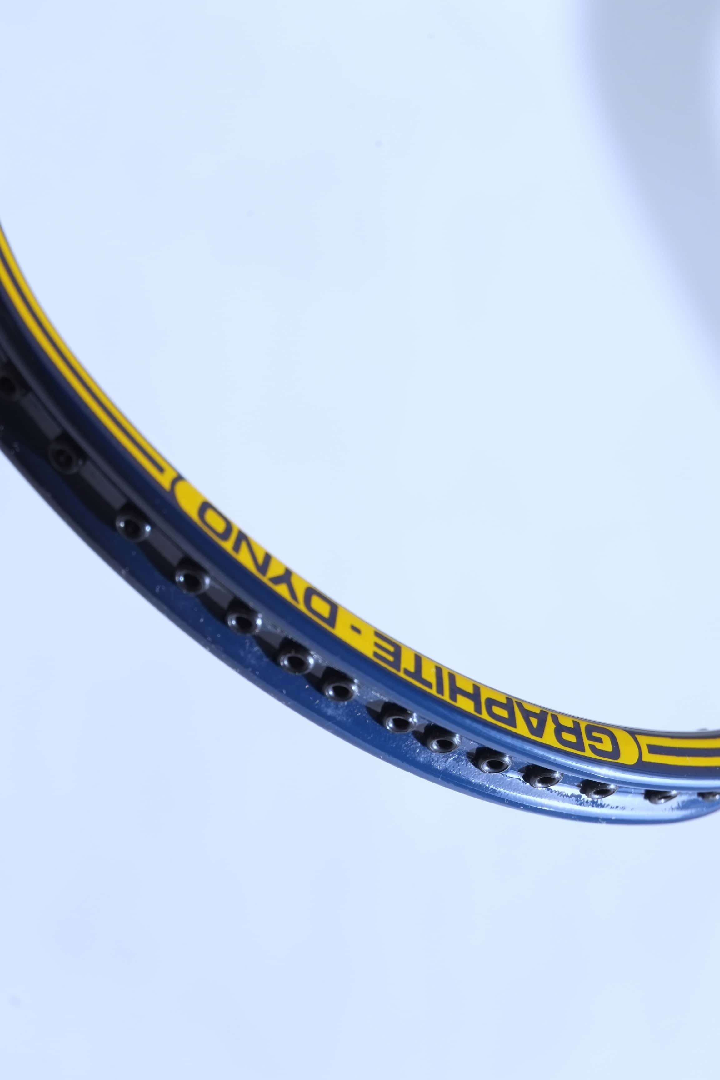 Macro side profile shot of the blue racquet frame highlighting a row of empty black string grommets along the outer channel.