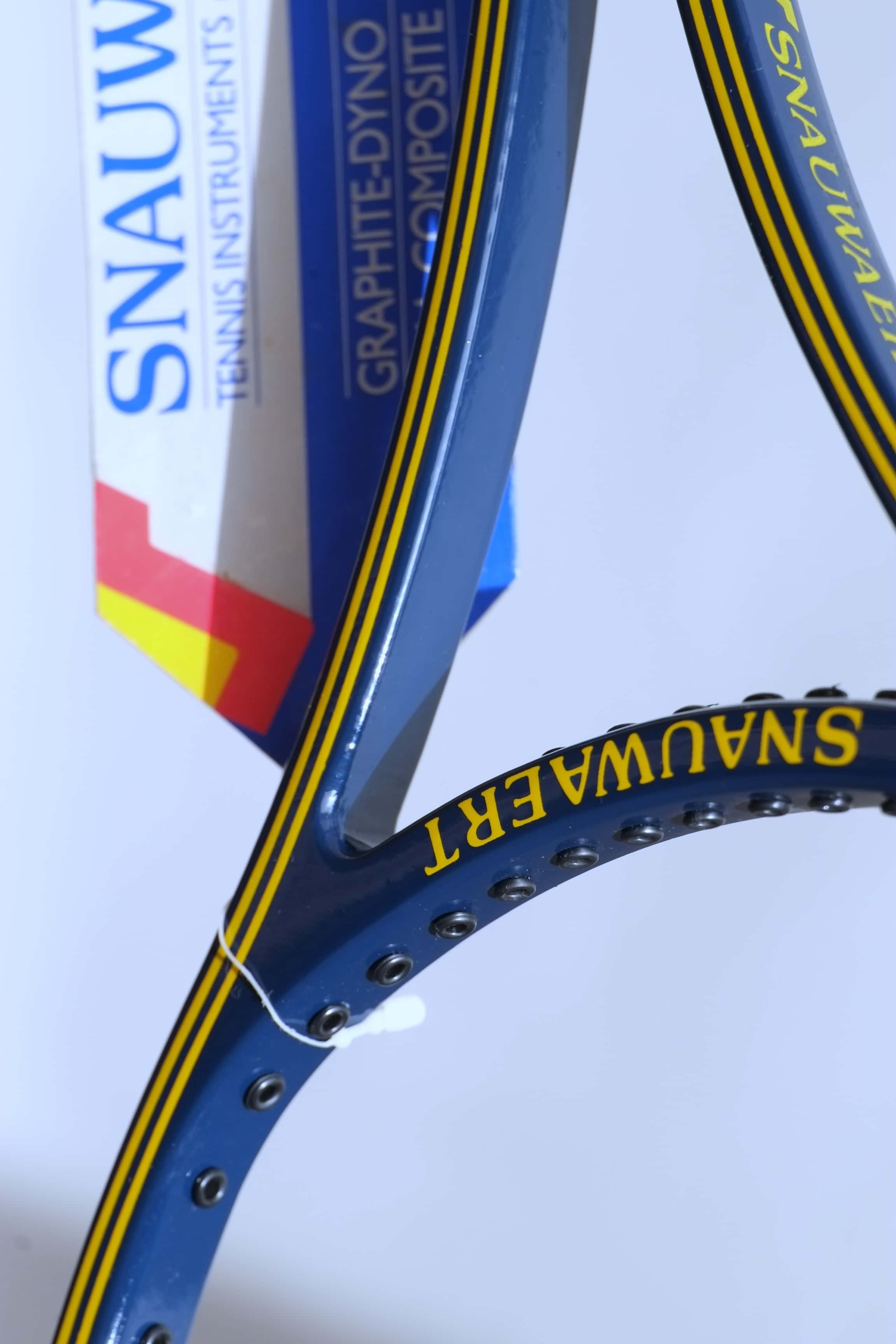 Close-up detail of the inner racquet throat and shaft, showing the yellow Snauwaert branding and the text on the vintage paper warranty hang tag.