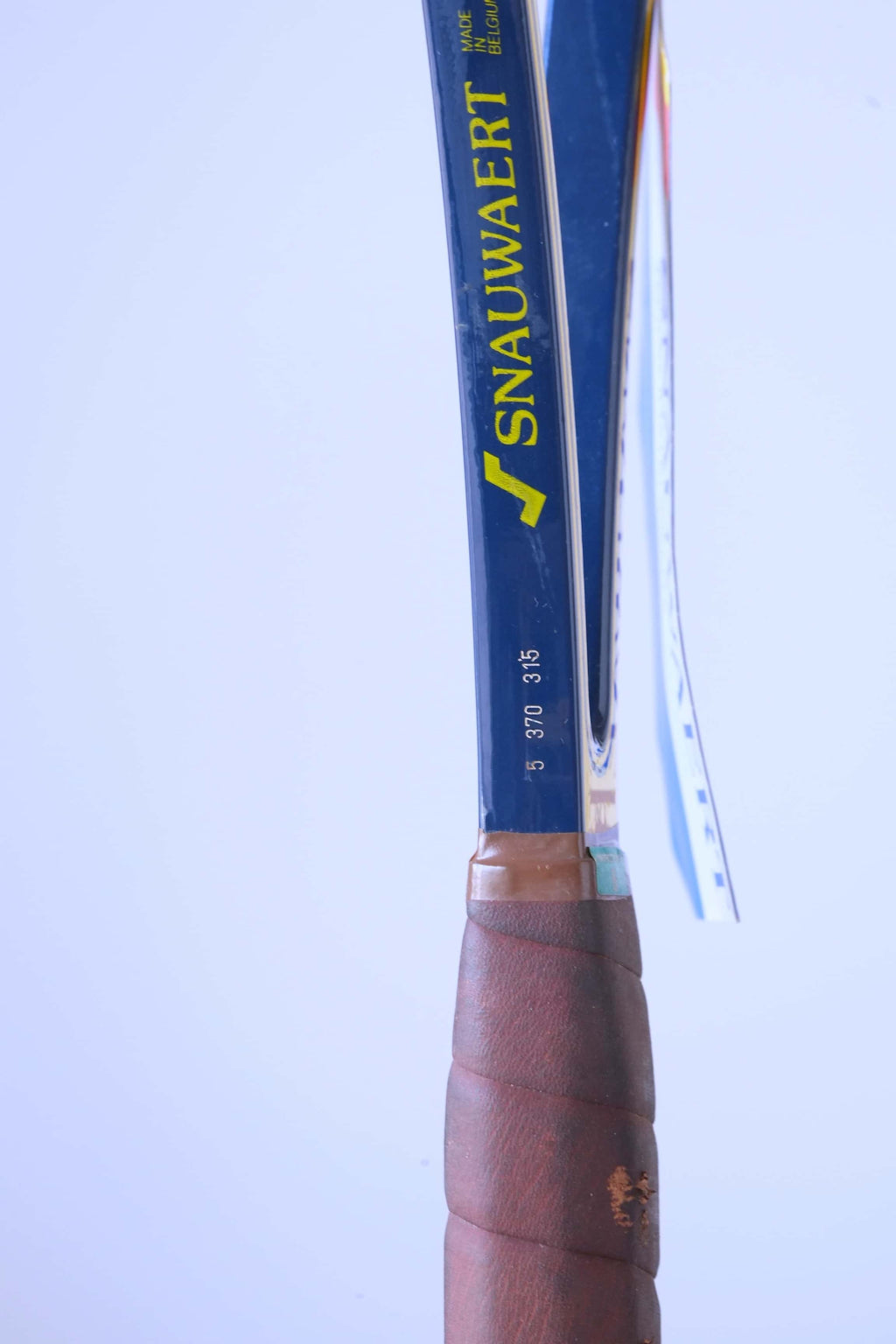 Close-up of the racquet handle's upper shaft featuring the yellow Snauwaert logo, "MADE IN BELGIUM" text, and printed grip/weight specifications above the leather wrap.