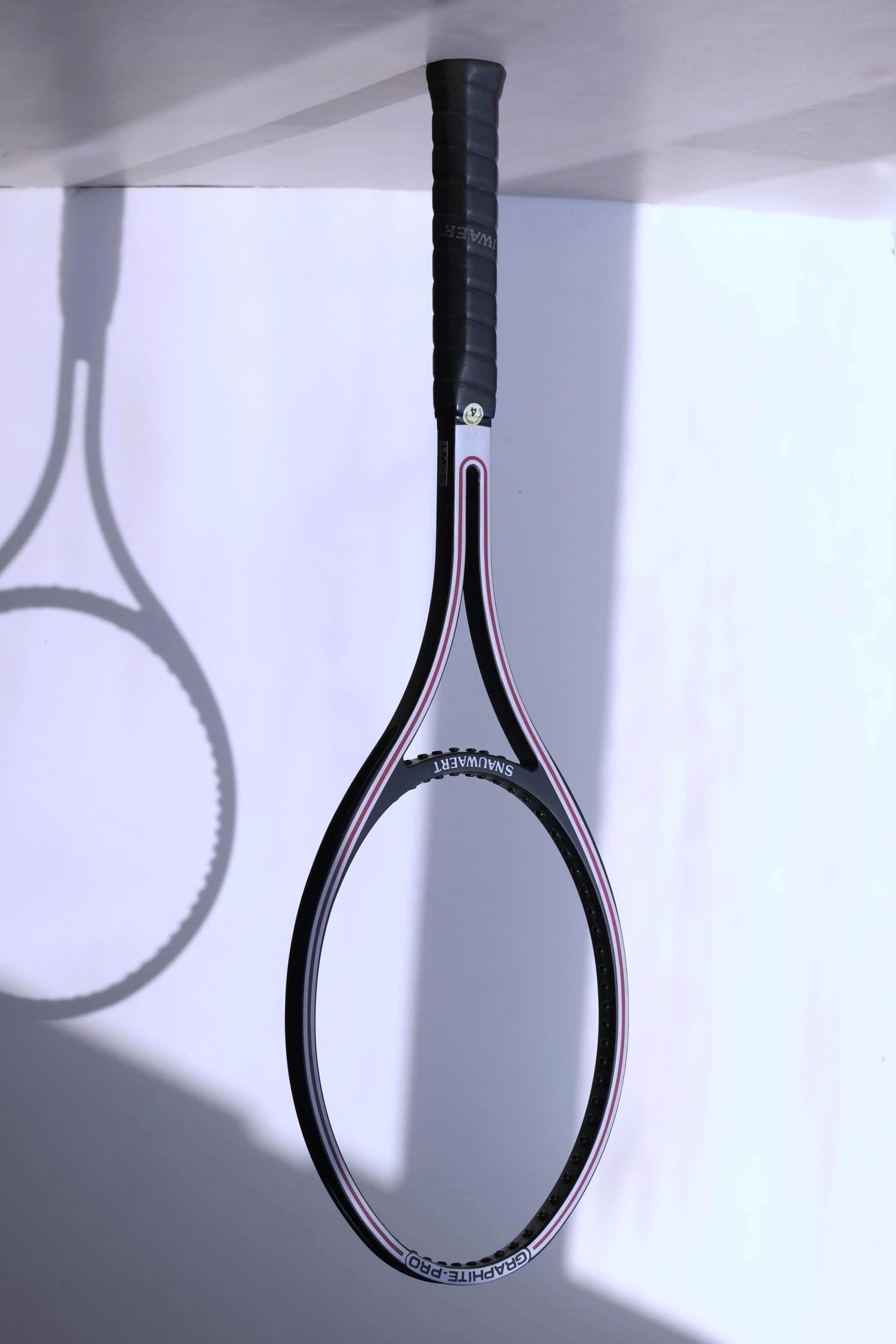 An unstrung vintage Snauwaert Graphite Pro tennis racquet stands vertically, casting a distinct shadow on a white background.