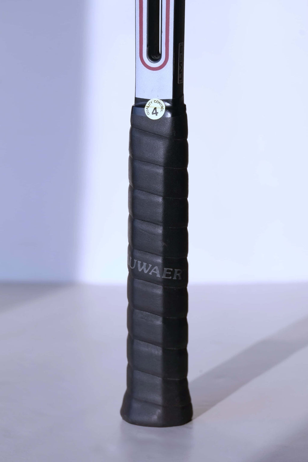 Detail of the racquet handle wrapped in a black textured grip with a round "Quality Control 4" sticker at the top.