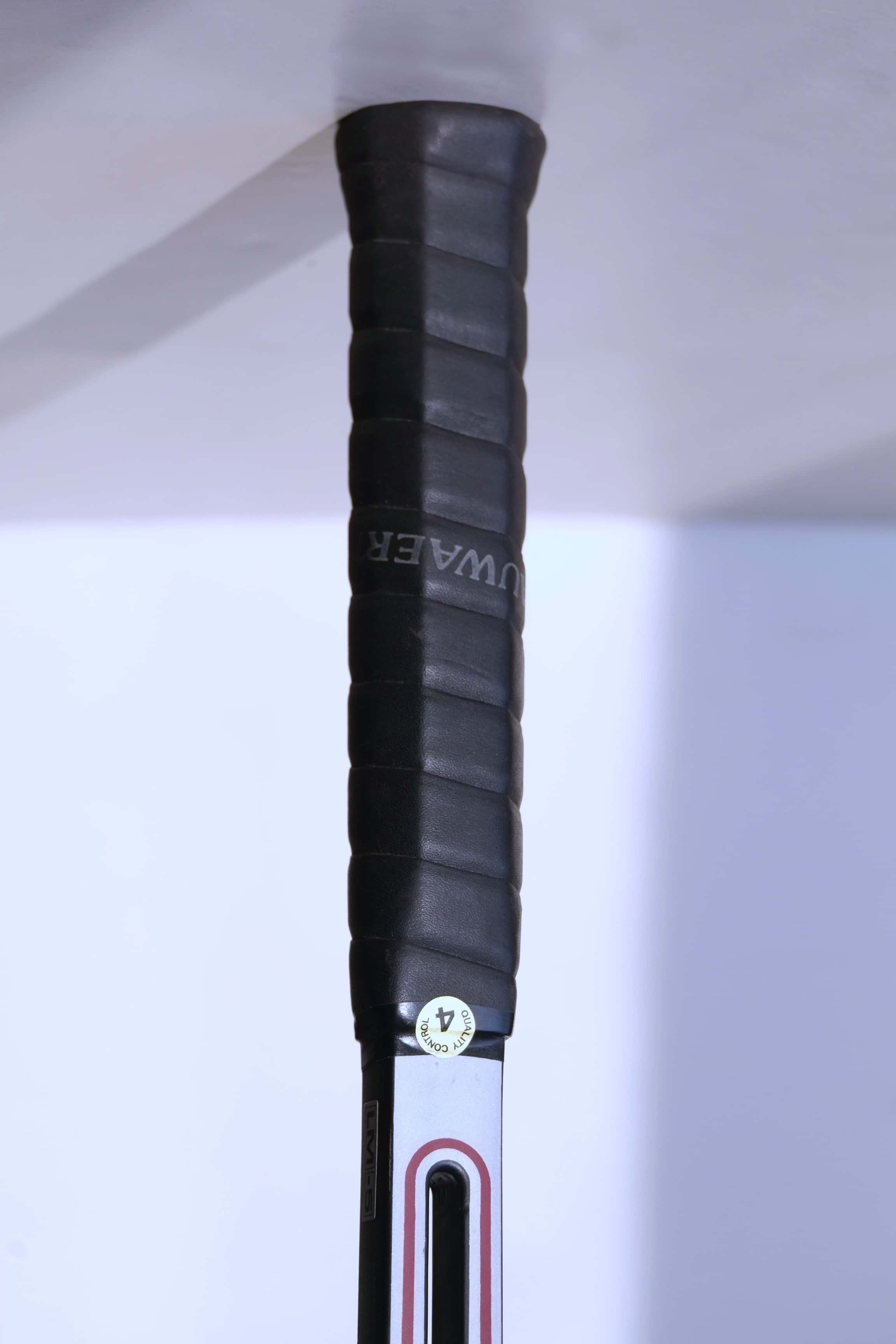 Detail of the racquet handle wrapped in a black textured grip with a round "Quality Control 4" sticker at the top.