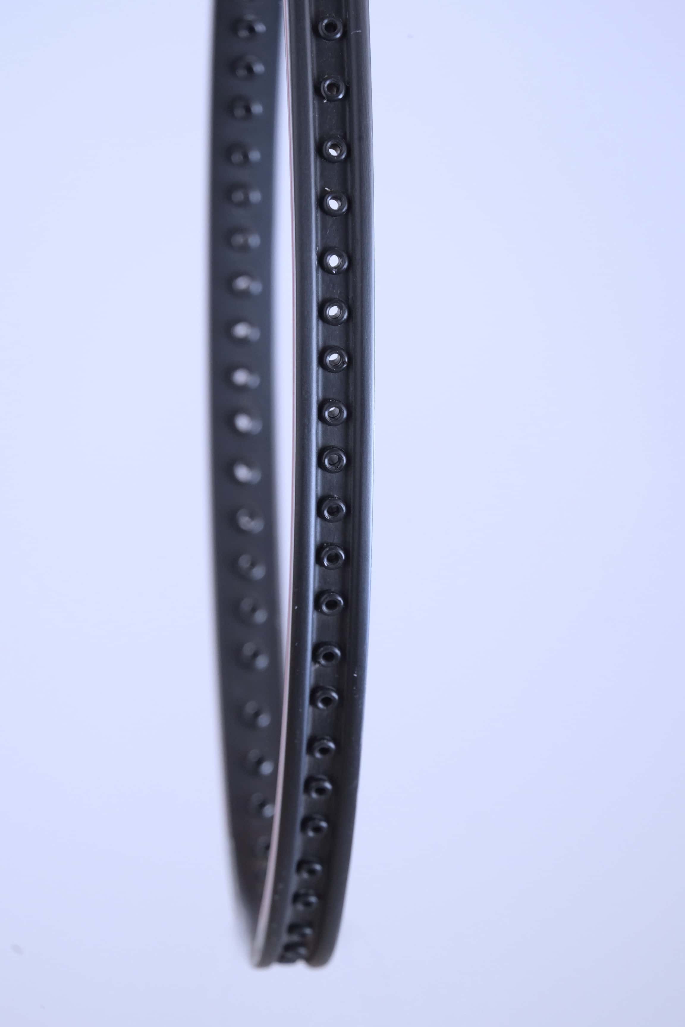 Profile view of the empty black racquet frame showing the precise row of grommet holes for stringing.