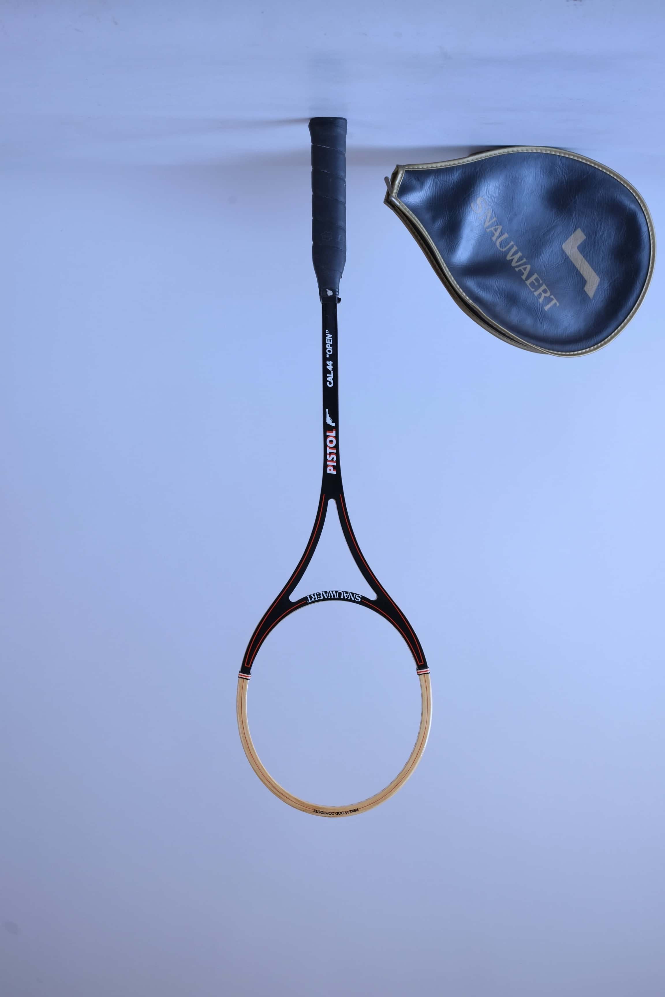 Vintage unstrung Snauwaert Pistol wood squash racket standing upright next to its matching black vinyl head cover.
