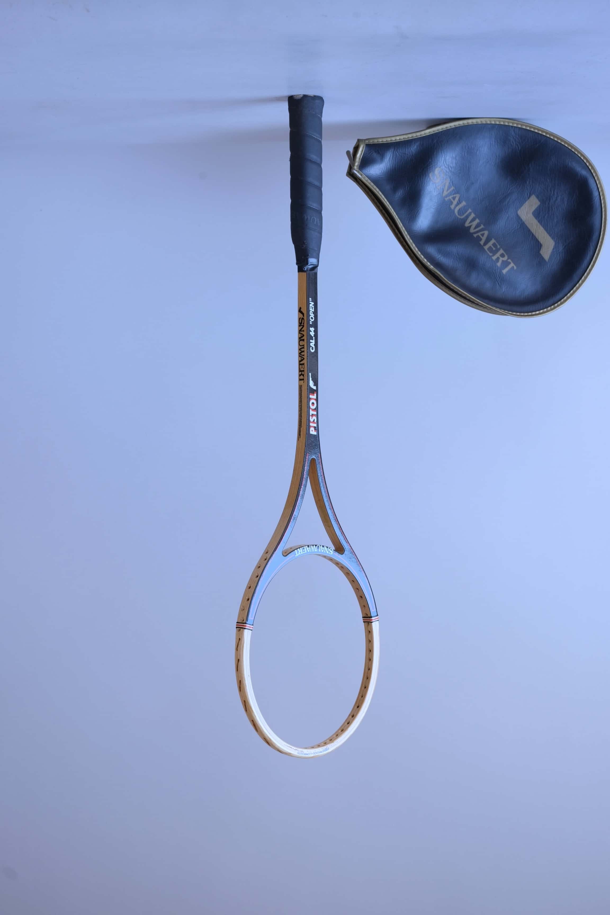 Vintage unstrung Snauwaert Pistol wood squash racket standing upright at an angle next to its matching black vinyl head cover.
