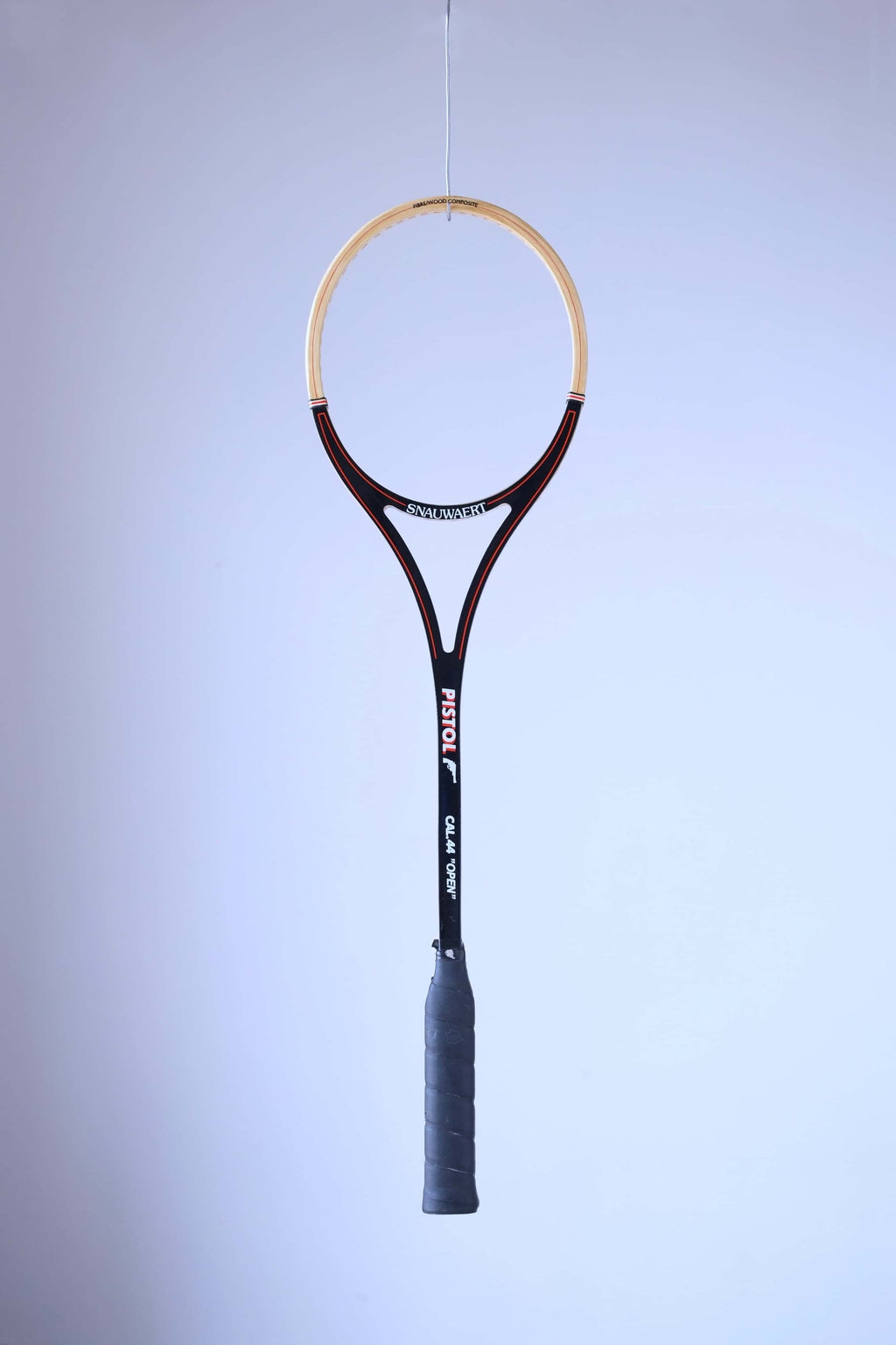 Full front view of an unstrung vintage Snauwaert Pistol Cal 44 Open squash racket hanging vertically against a plain background.