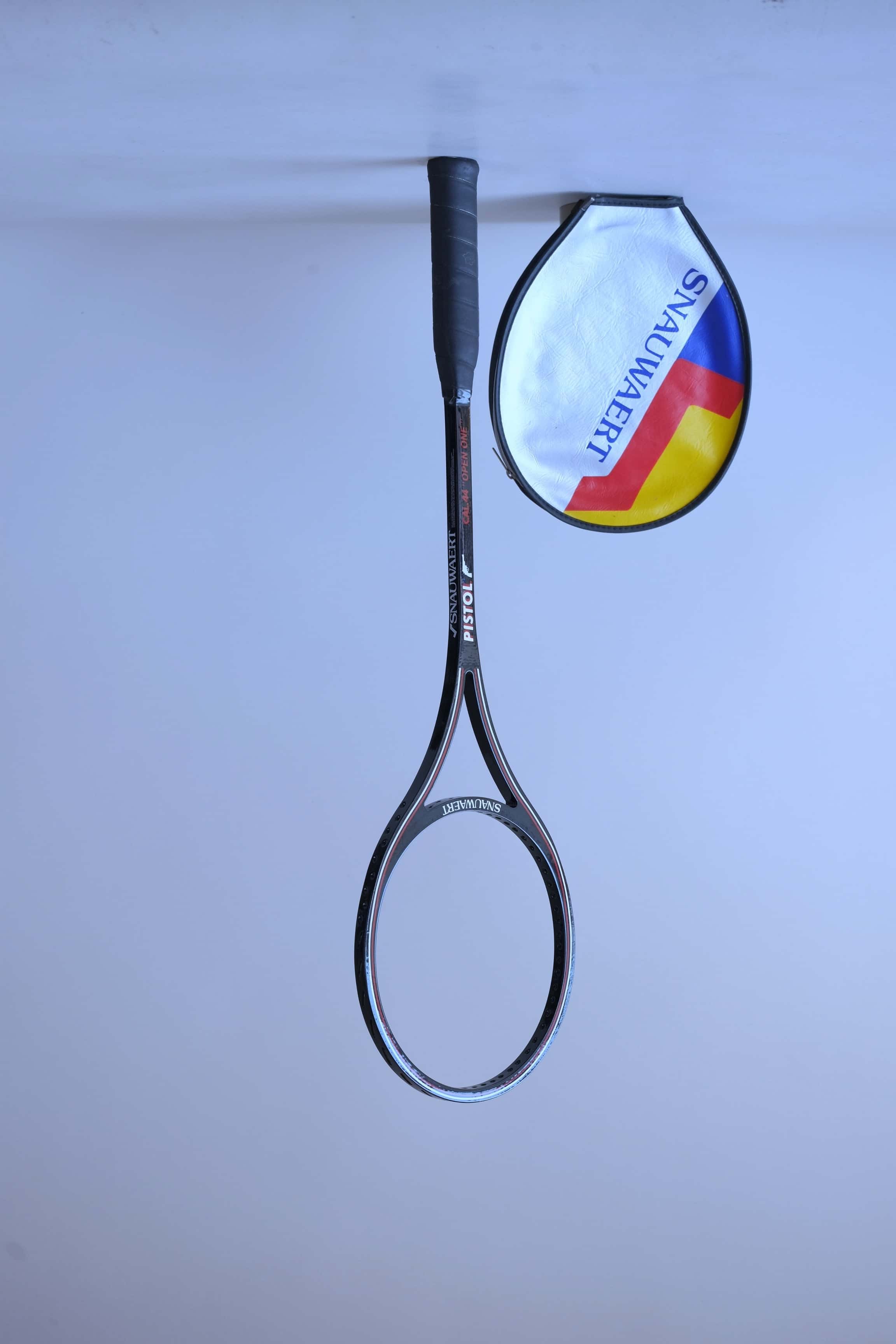 Angled view of an unstrung black vintage Snauwaert Pistol squash racket standing next to its vinyl cover.