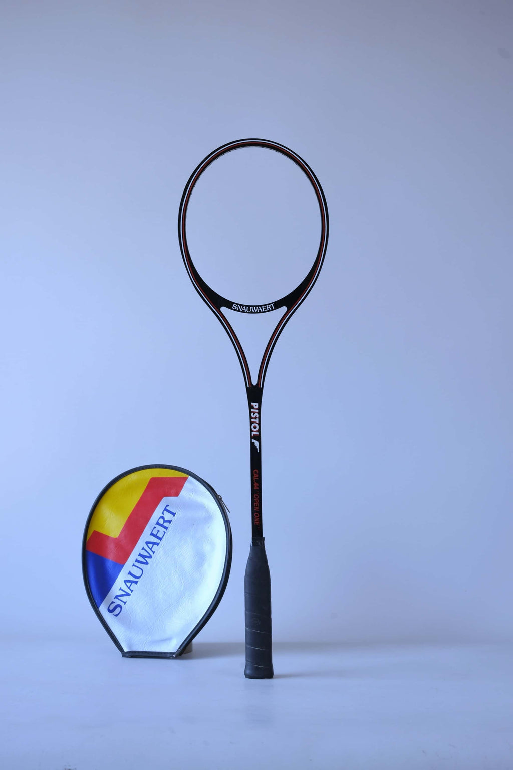 Vintage unstrung black graphite Snauwaert Pistol squash racket standing upright next to a grey vinyl head cover with yellow and blue stripes.