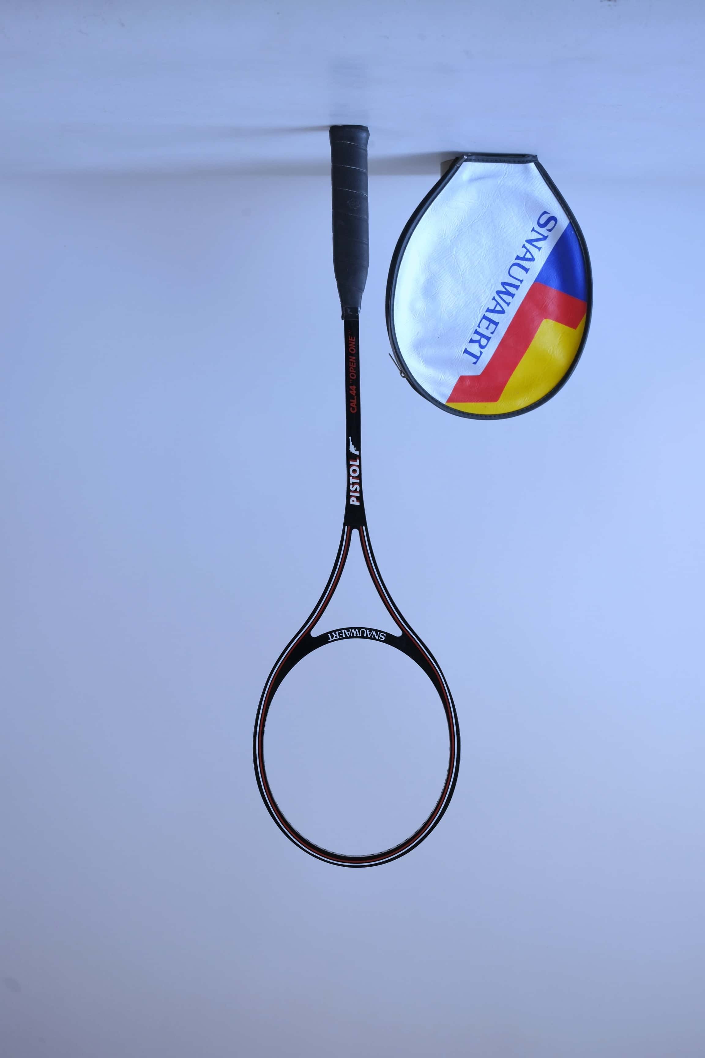 Vintage unstrung black graphite Snauwaert Pistol squash racket standing upright next to a grey vinyl head cover with yellow and blue stripes.