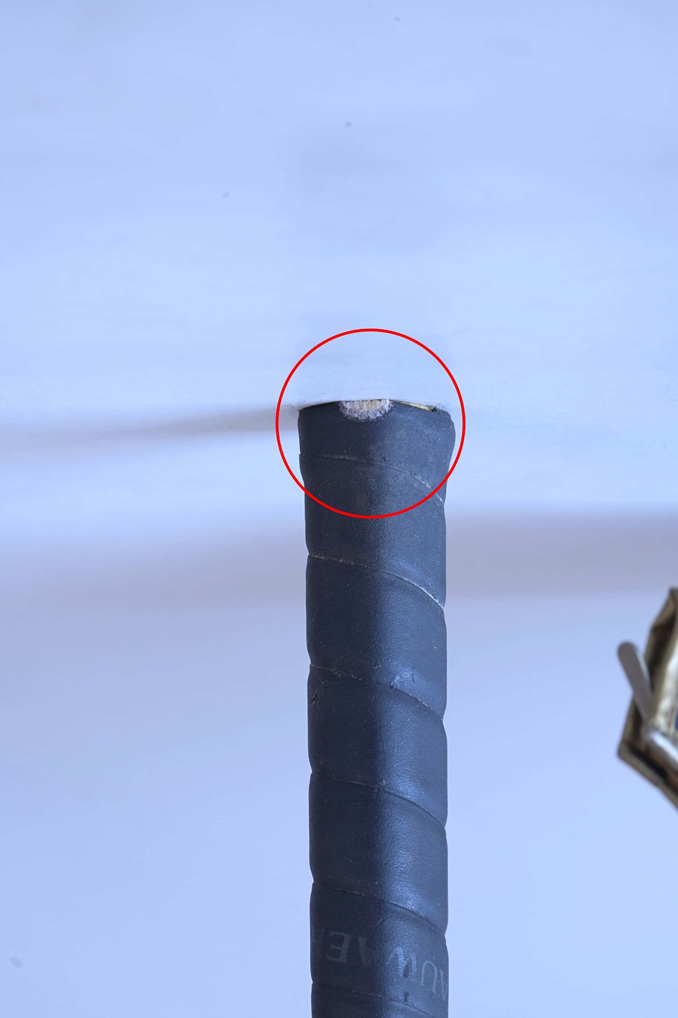 Close-up of a squash racket handle with a red circle highlighting a small nick of wear at the very bottom edge of the black leather grip.