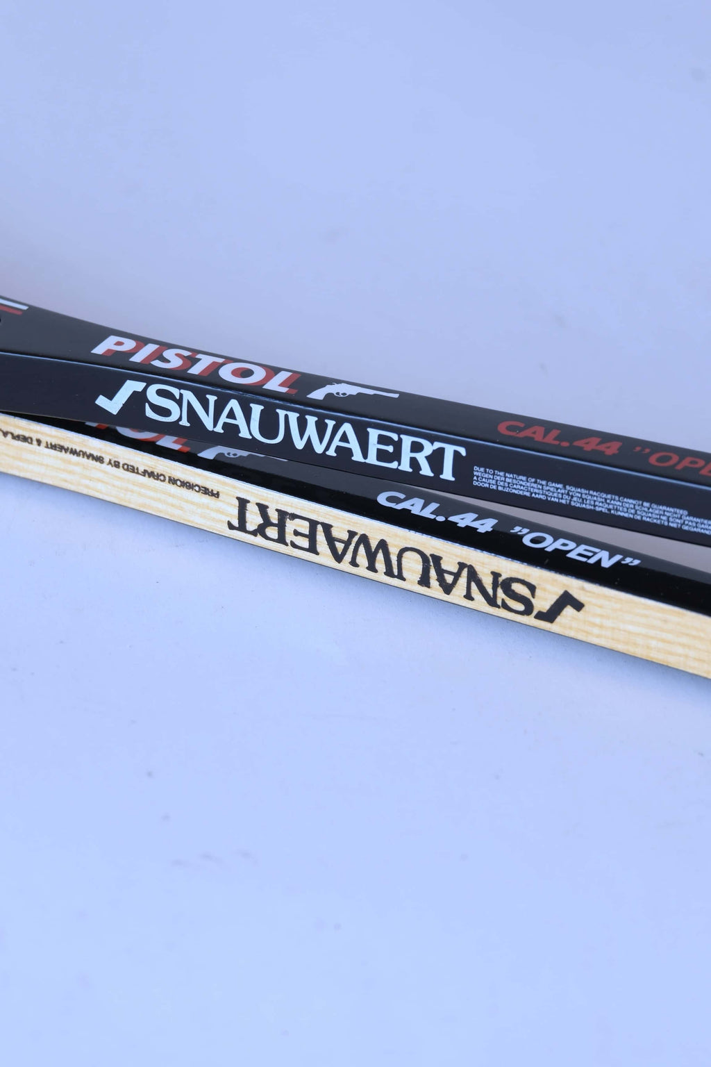 Side-by-side comparison view of the shafts of a glossy black graphite racket and a natural wood grain racket, both displaying Snauwaert branding.