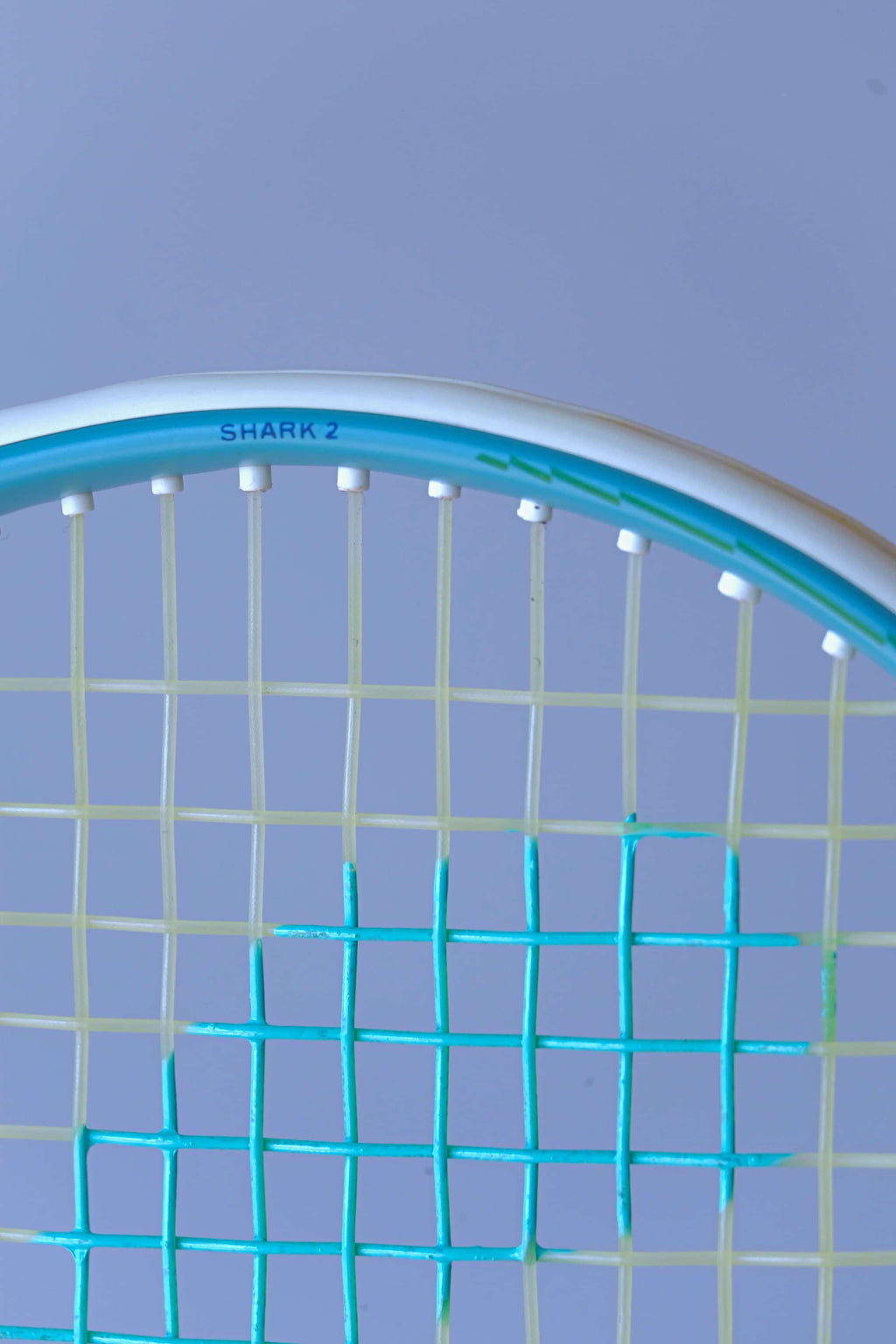 Close-up of the Snauwaert Shark 2 squash racquet head showing white grommets, teal and yellow-green string pattern and Shark 2 model name printed on the frame.