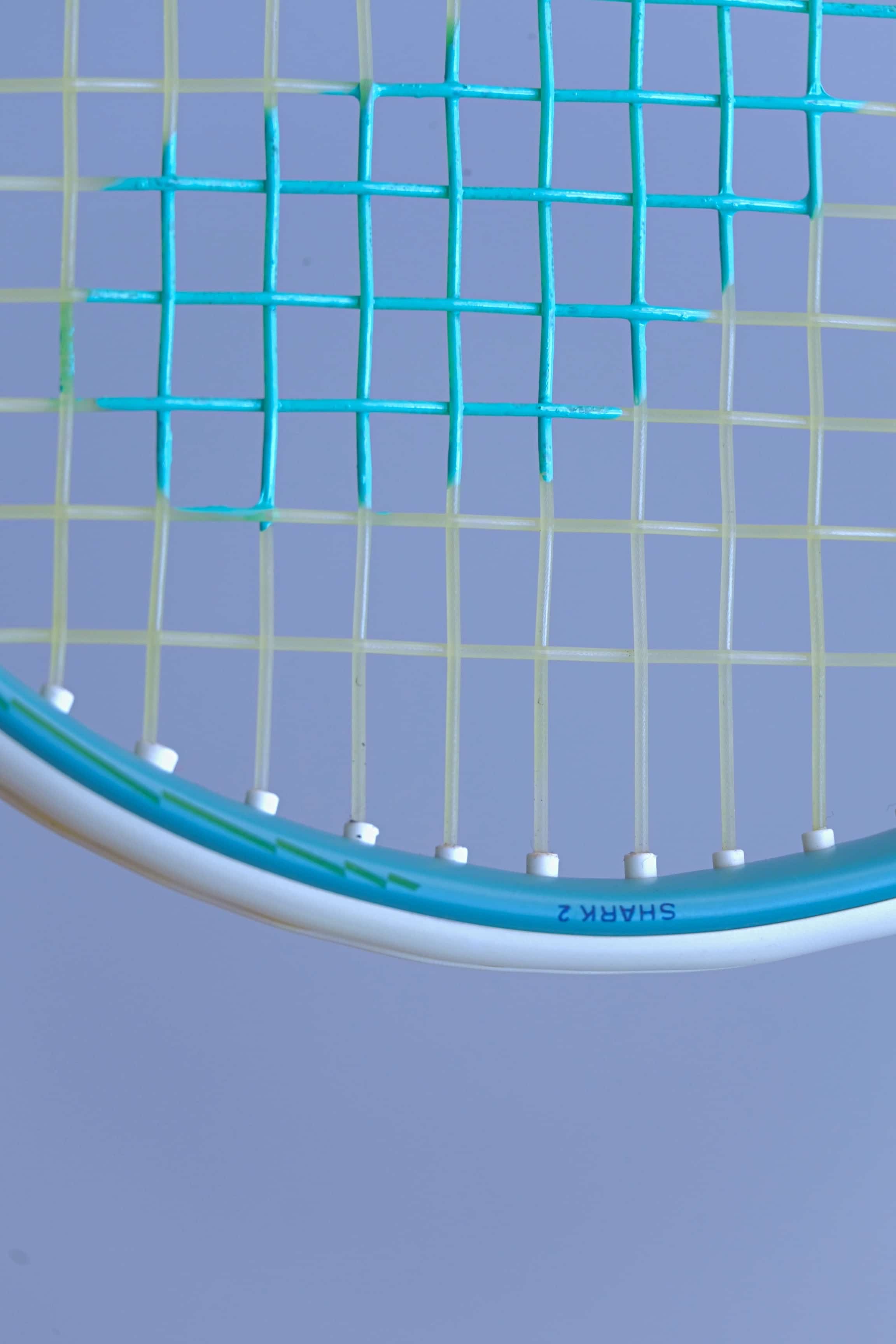 Close-up of the Snauwaert Shark 2 squash racquet head showing white grommets, teal and yellow-green string pattern and Shark 2 model name printed on the frame.
