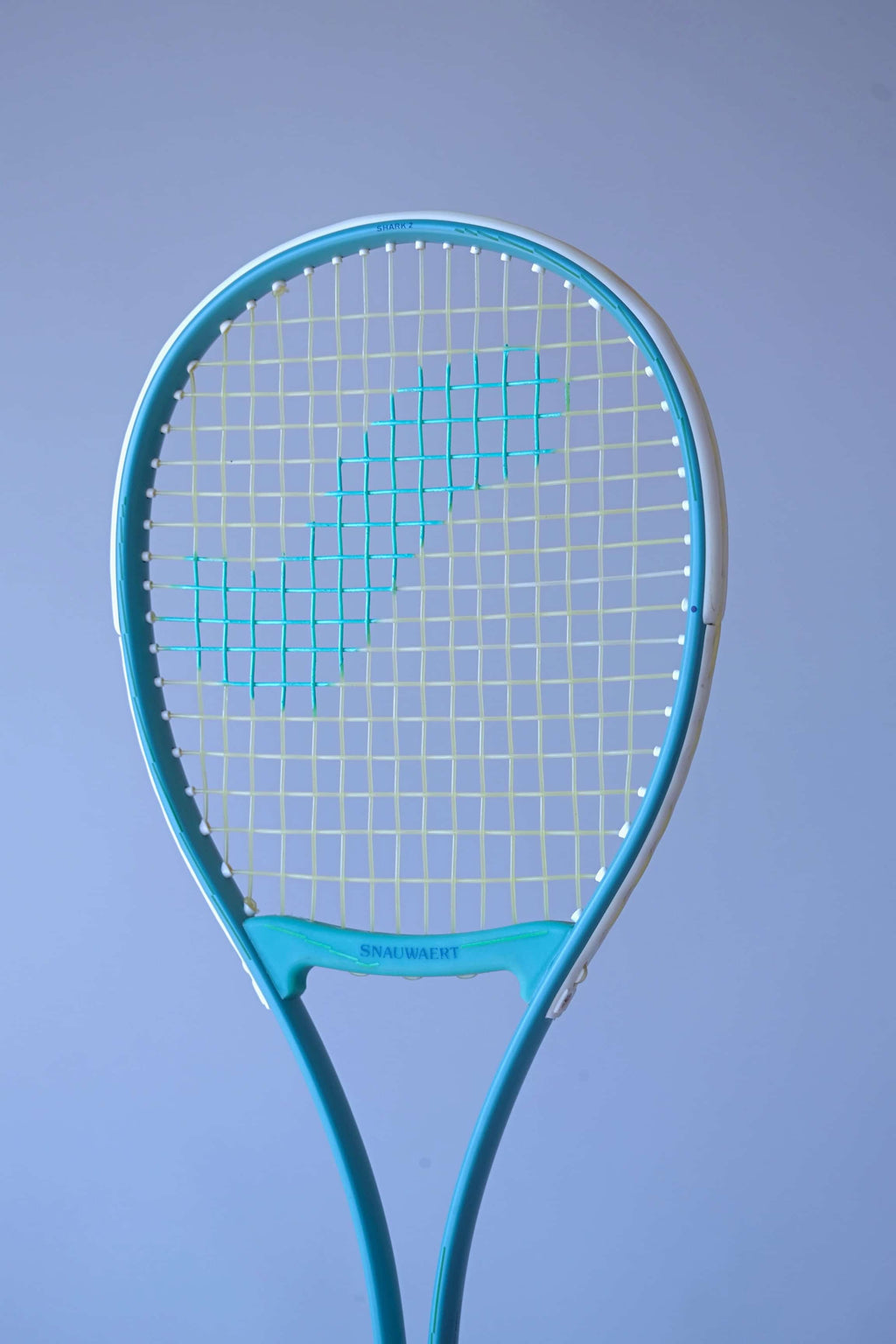 Angled front view of the Snauwaert Shark 2 squash racquet head showing teal and white frame, Snauwaert throat bridge and teal cross-string pattern on yellow-green mains.