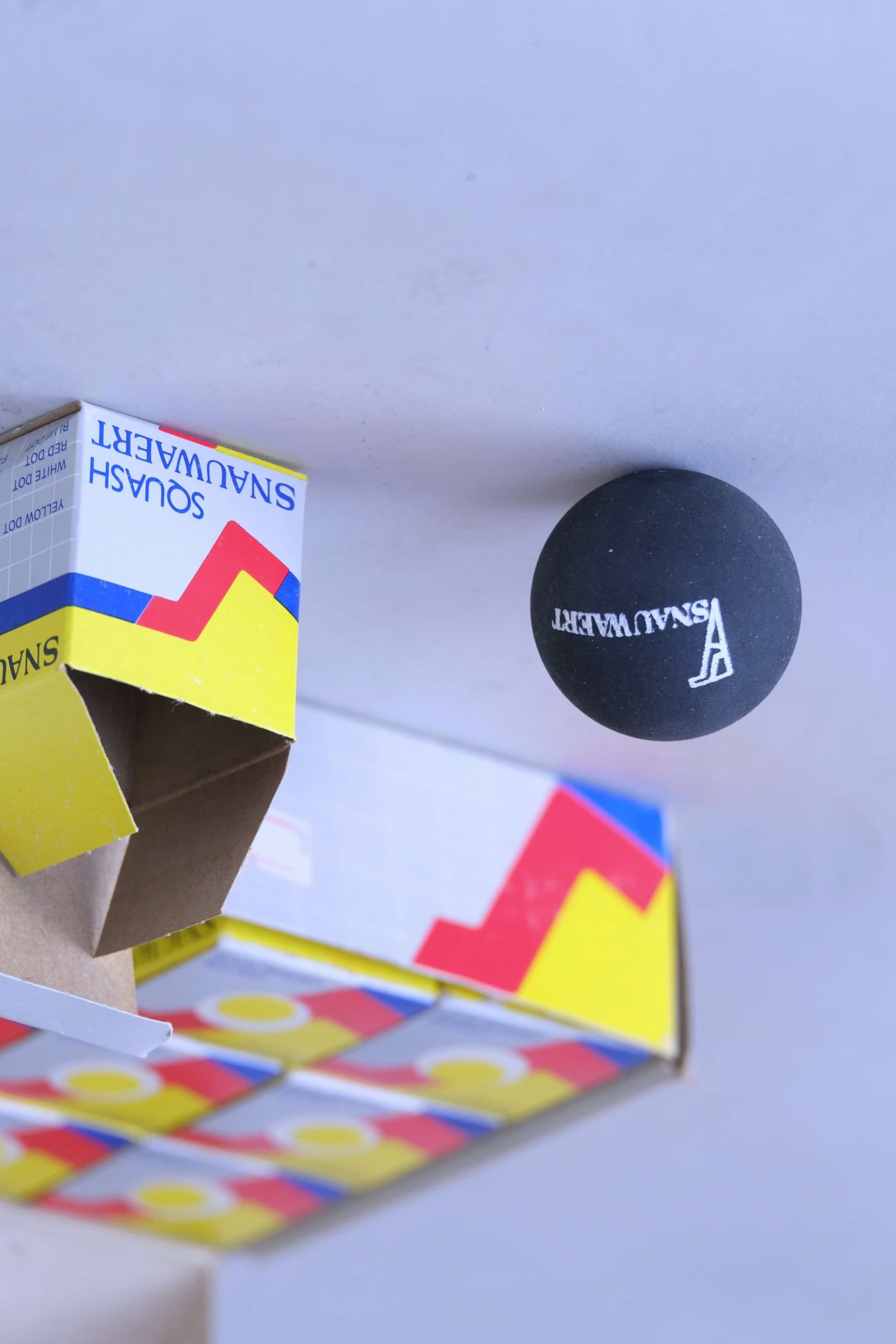 Close-up of a vintage black rubber squash ball featuring a printed Snauwaert logo.